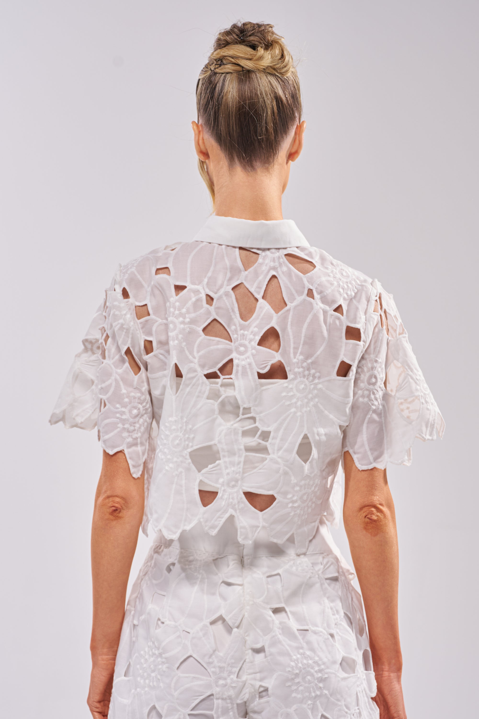 A woman with blonde hair in an updo, seen from behind, wears the DOLLY FLORAL BUTTON DOWN IN WHITE with matching bottoms—perfect summer resort wear—featuring large cutouts and intricate embroidery against a plain background. 3