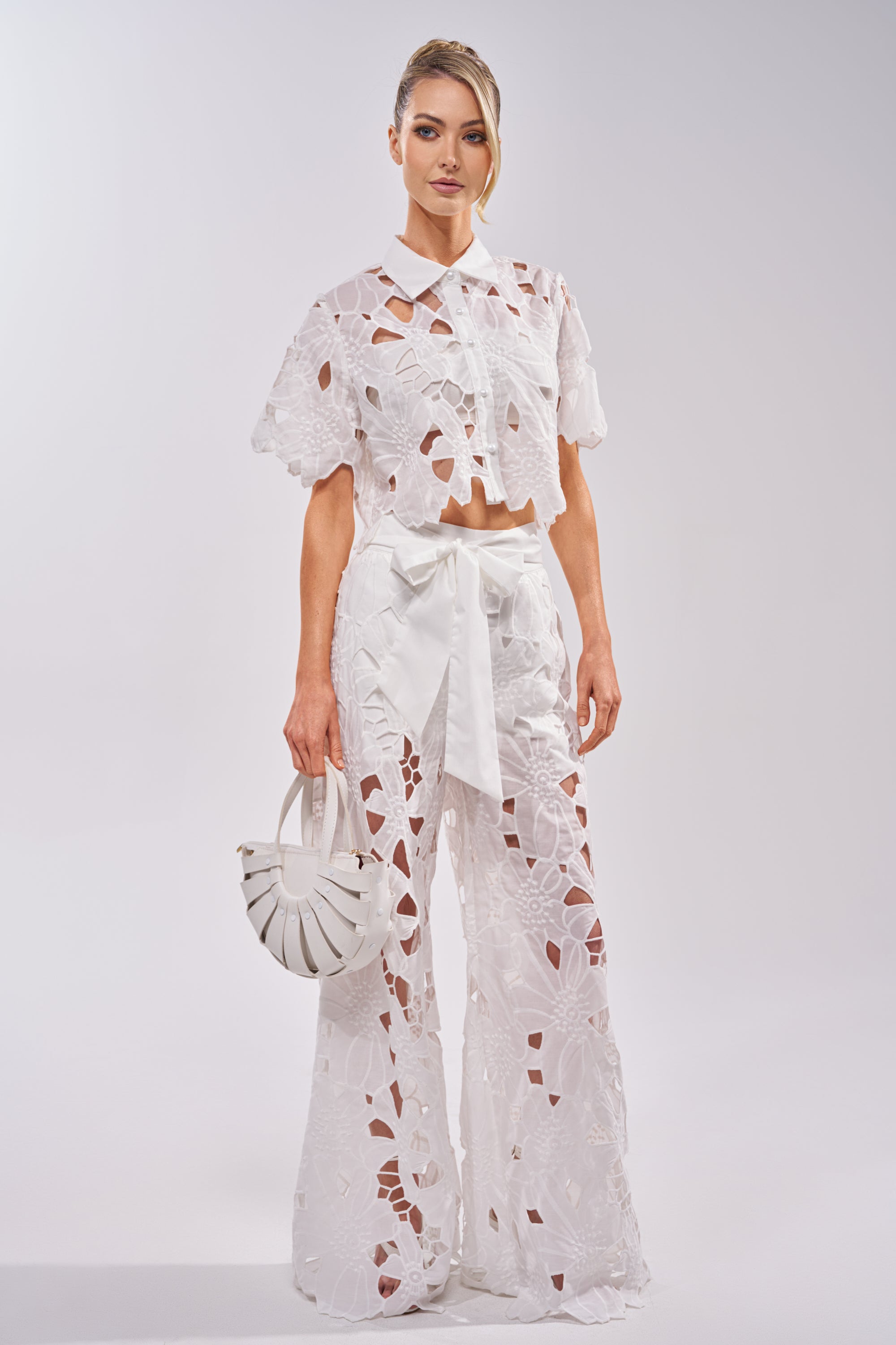 A woman stands against a plain background wearing the DOLLY FLORAL BUTTON DOWN IN WHITE, a matching cropped white floral lace shirt with wide-leg cut-out pants. She carries a small white round handbag, her hair styled to the side. 4
