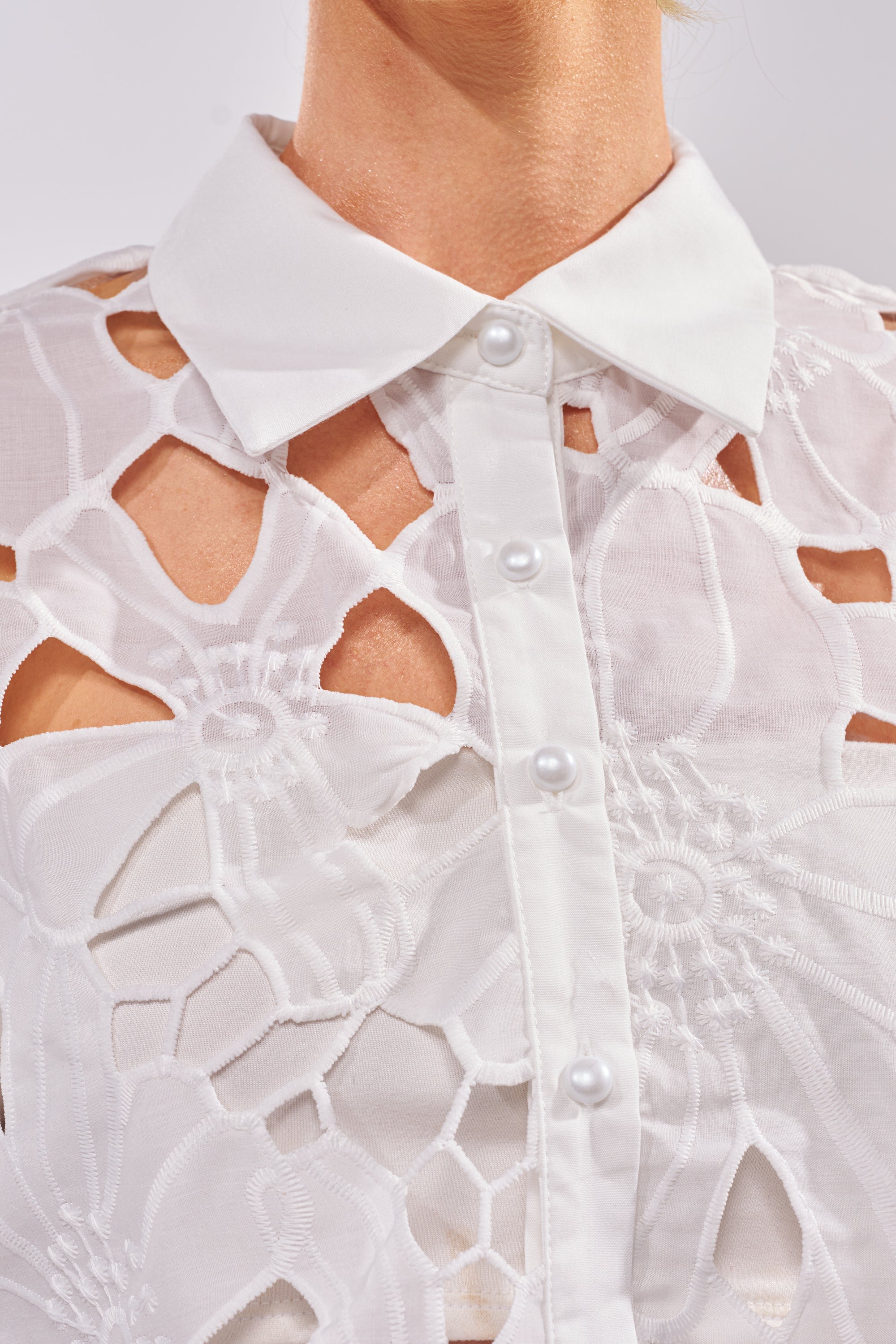 Close-up of the DOLLY FLORAL BUTTON DOWN IN WHITE, a cropped lace top with pearl-like buttons and delicate cut-out floral patterns—ideal for summer resort wear. The image highlights the neck and upper chest. 5