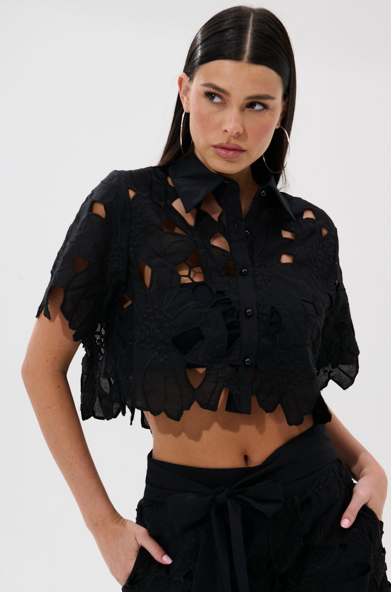 A woman with long dark hair wears the DOLLY FLORAL BUTTON DOWN BLOUSE, a black, short-sleeve cropped lace top with cutout patterns, paired with matching high-waisted shorts. She looks to the side with one hand on her hip. 0