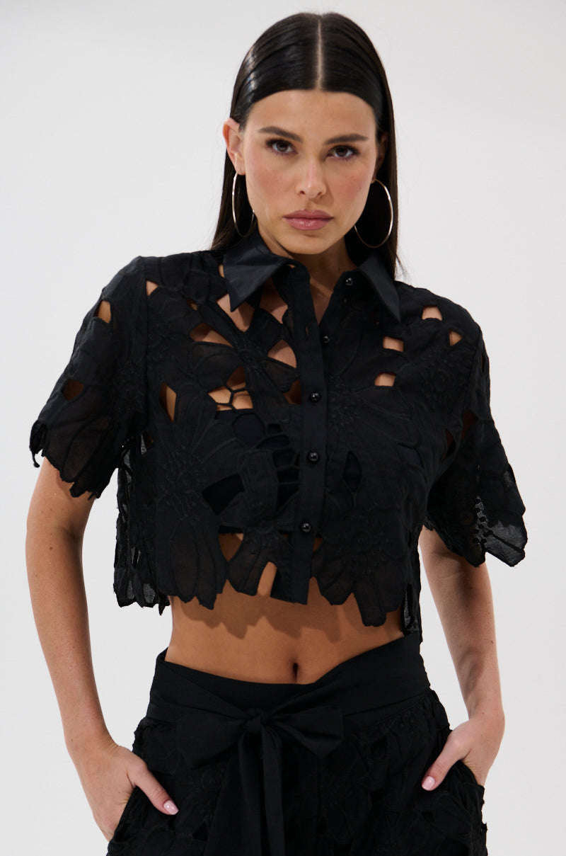 A woman with straight dark hair and hoop earrings models the DOLLY FLORAL BUTTON DOWN BLOUSE—black, cropped, and boxy with floral cutouts—paired with matching high-waisted bottoms. She poses confidently against a light background. 1
