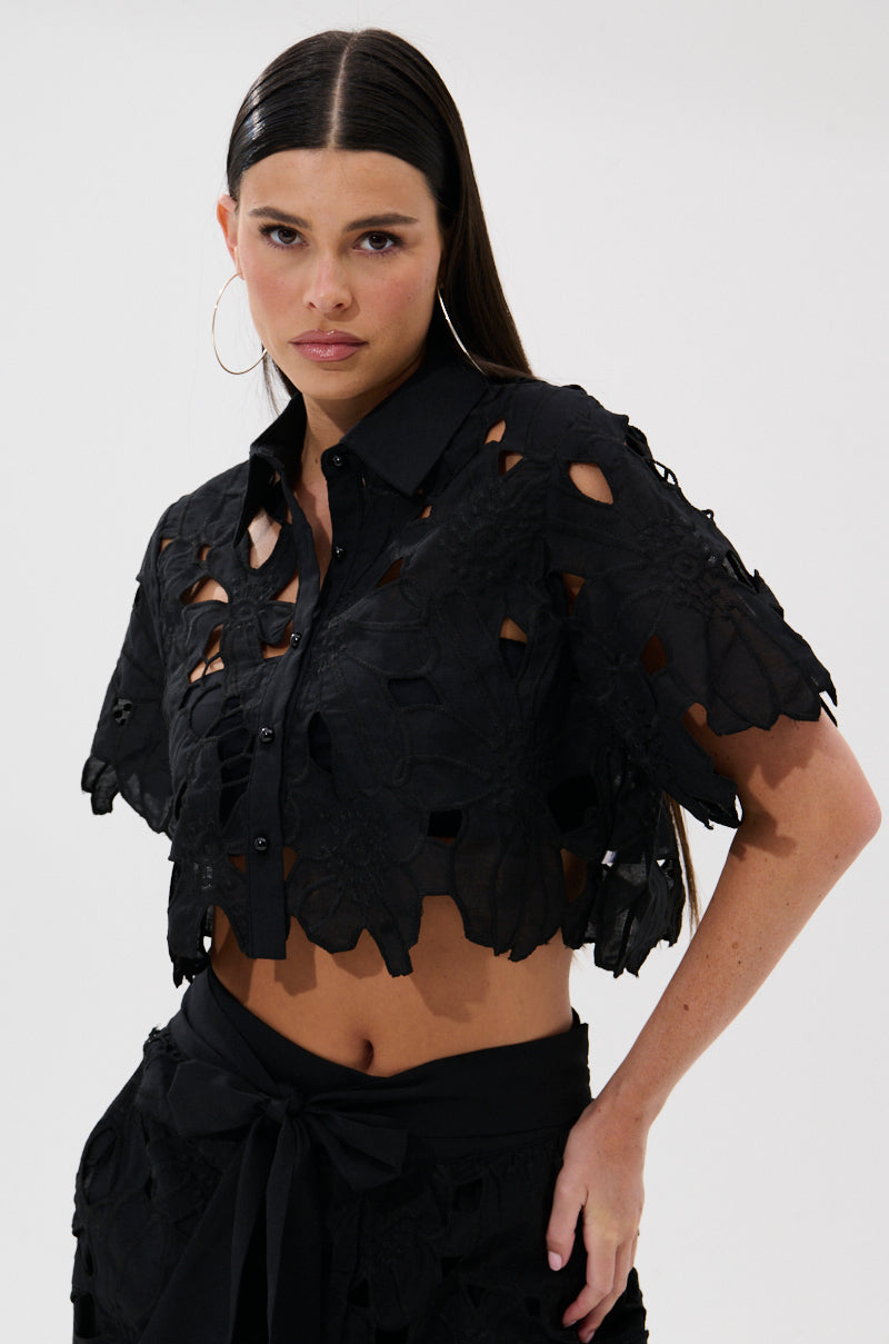 A woman with straight dark hair and hoop earrings wears the DOLLY FLORAL BUTTON DOWN BLOUSE, a black, short-sleeved, cropped lace top, styled with a matching skirt against a plain white background. 2