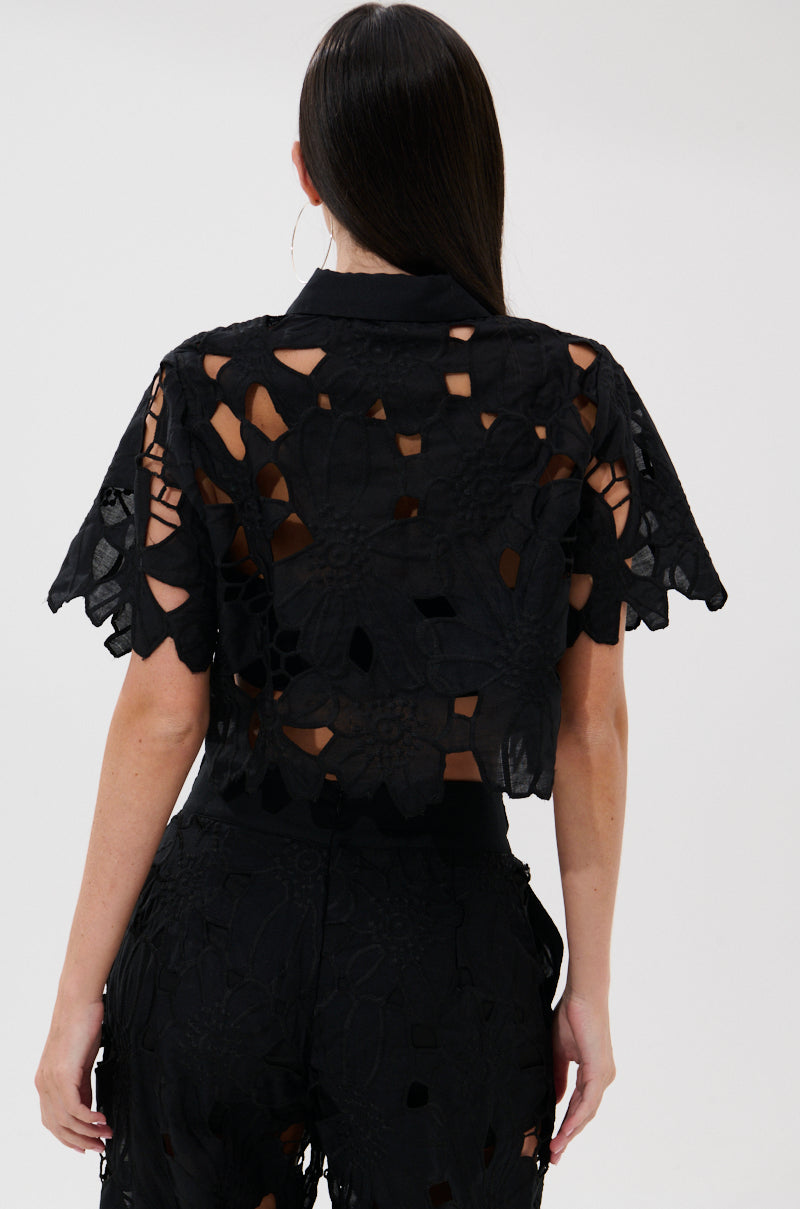 A person with long, straight dark hair is shown from the back, wearing the DOLLY FLORAL BUTTON DOWN BLOUSE with large lace cutouts and matching high-waisted pants. The sheer, boxy top stands out against a plain white background. 4