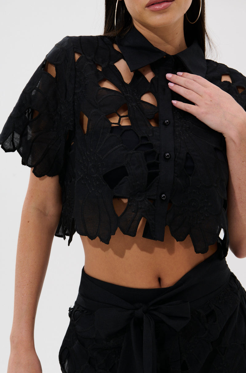 A woman wears the DOLLY FLORAL BUTTON DOWN BLOUSE—a black cropped top with large floral cutouts—and matching high-waisted, tie-waist shorts. Her right hand gently touches her chest. 8