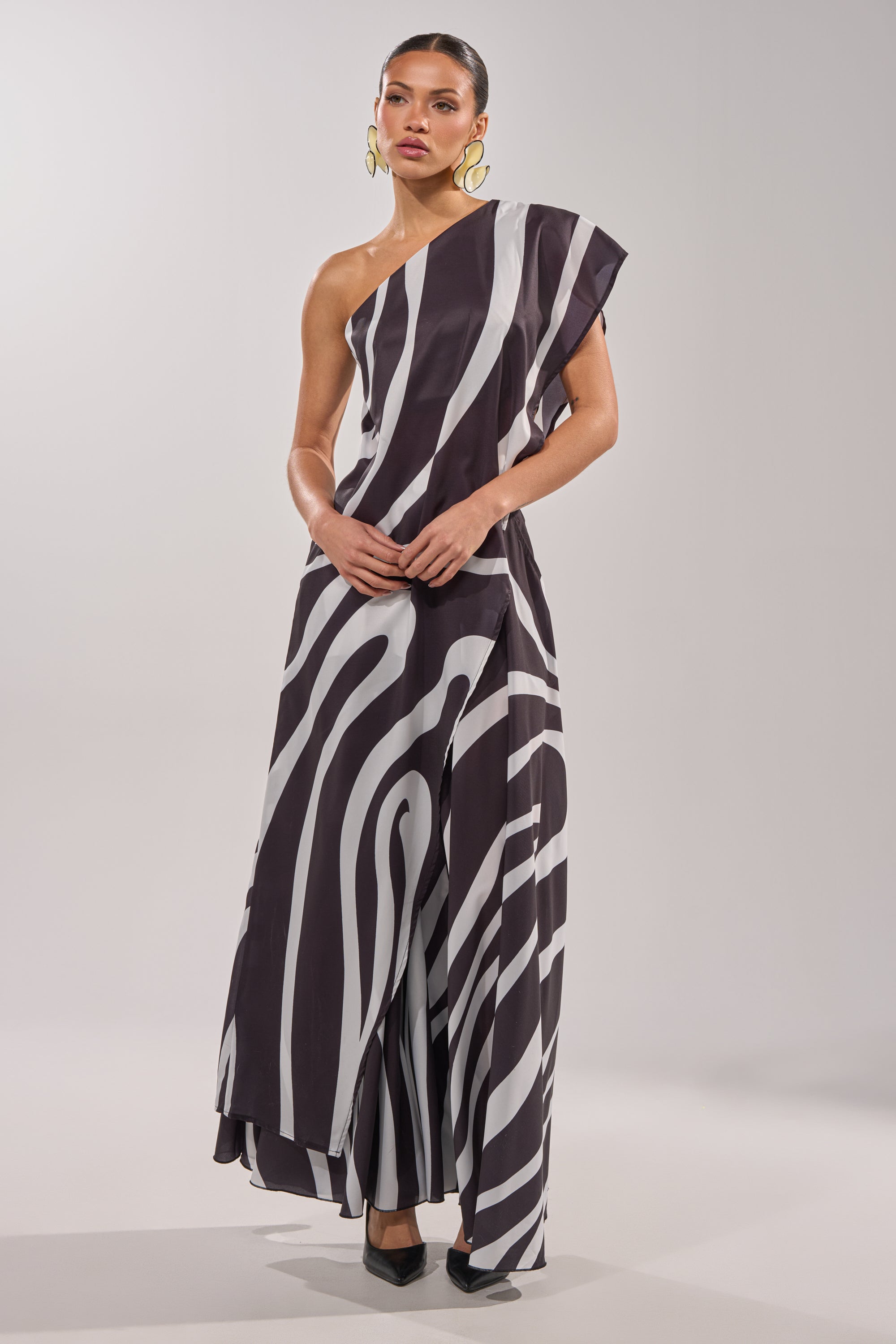 A woman wears the TRINA ELONGATED SINGLE SHOULDER TOP in a bold black and white swirl print, styled with slicked-back hair, large gold hoop earrings, and black pointed-toe heels against a plain background. 1