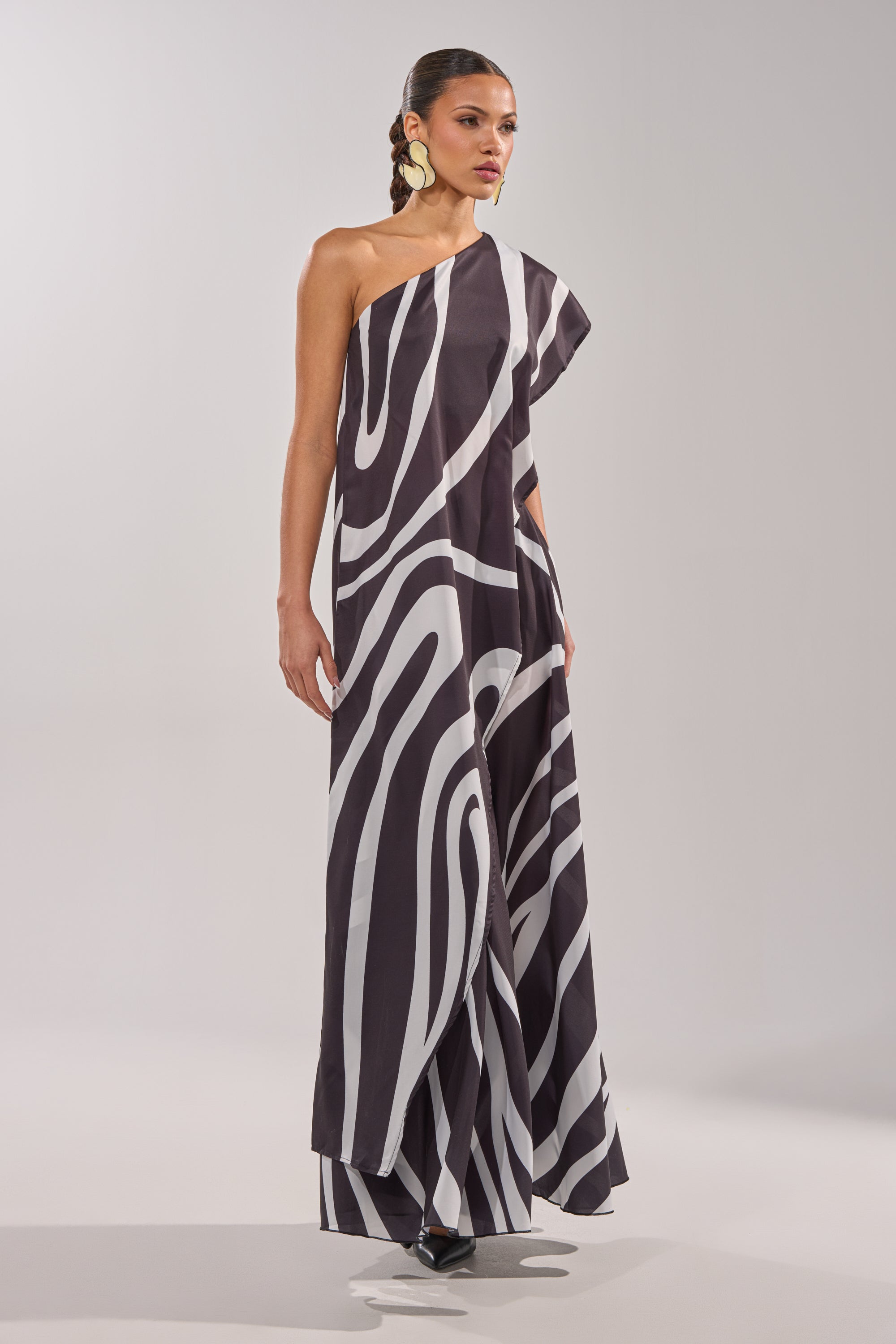 A woman poses confidently against a plain light background, wearing the TRINA ELONGATED SINGLE SHOULDER TOP styled as a floor-length dress with bold abstract stripes. Her hair is pulled back and she accessorizes with large earrings. 2