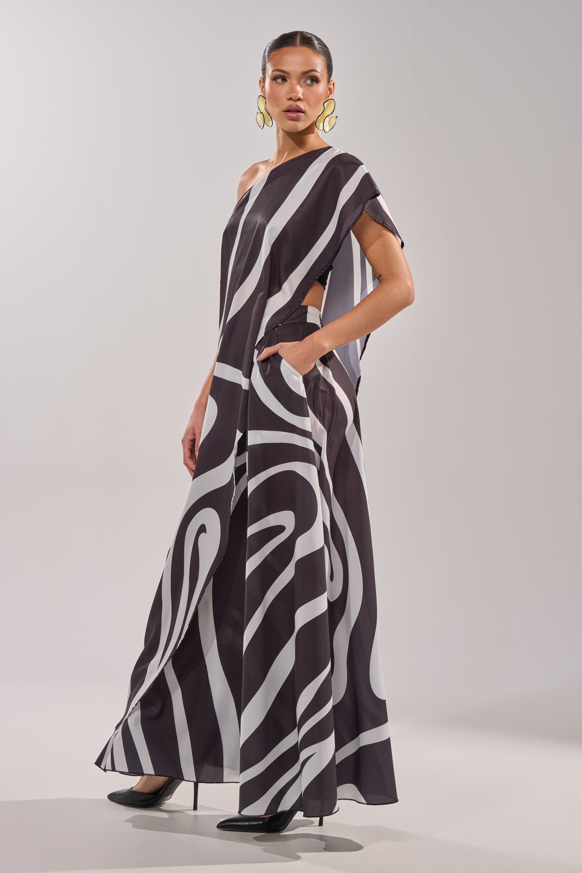 A woman models the TRINA ELONGATED SINGLE SHOULDER TOP, featuring a black and white swirl print and asymmetrical design. She pairs it with gold hoop earrings and black heels, making the bold top pop against a plain, light background. 3
