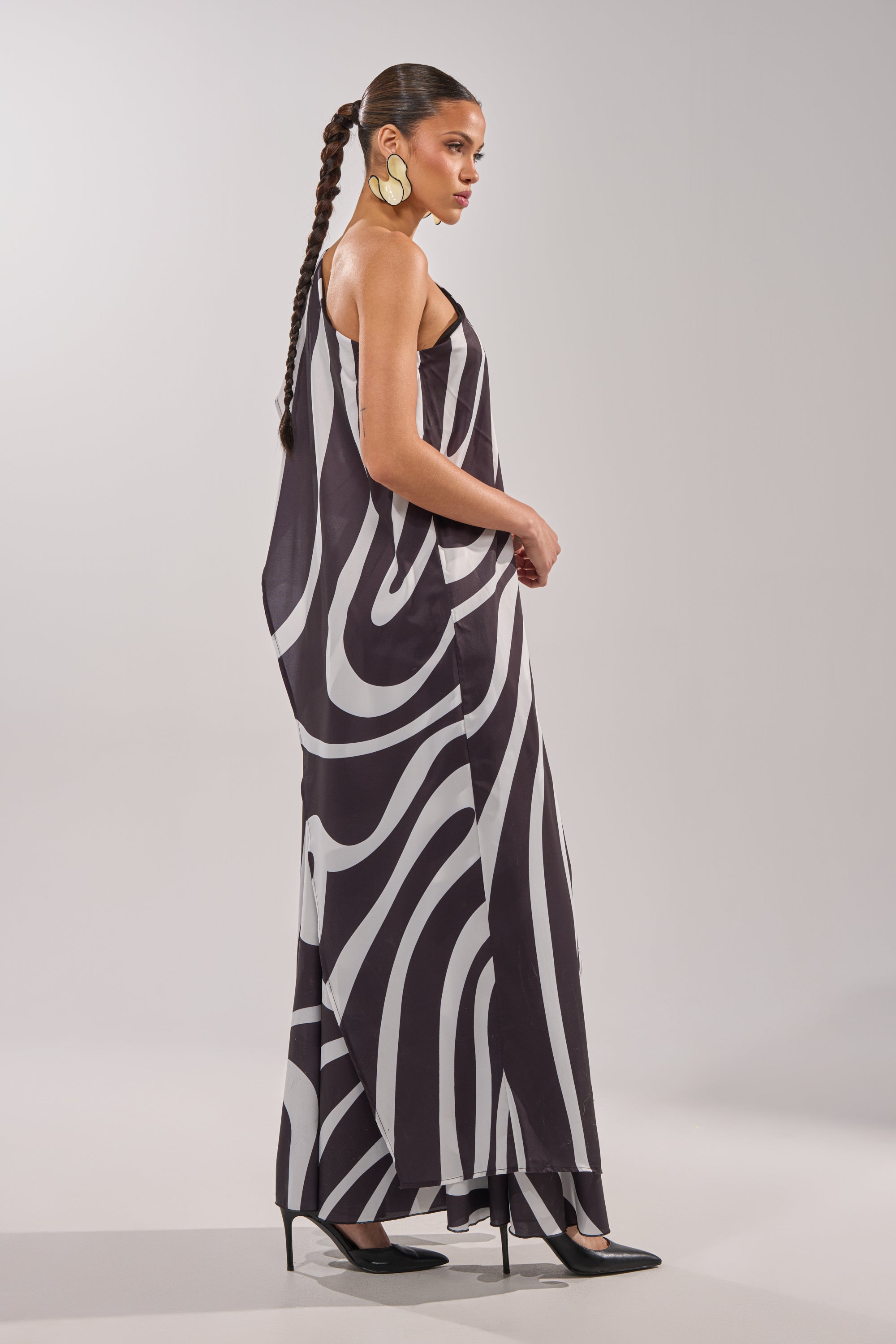 A woman with a long braid wears the TRINA ELONGATED SINGLE SHOULDER TOP, styled as a statement piece with black heels and large gold earrings, posing in profile against a plain light gray background. 4