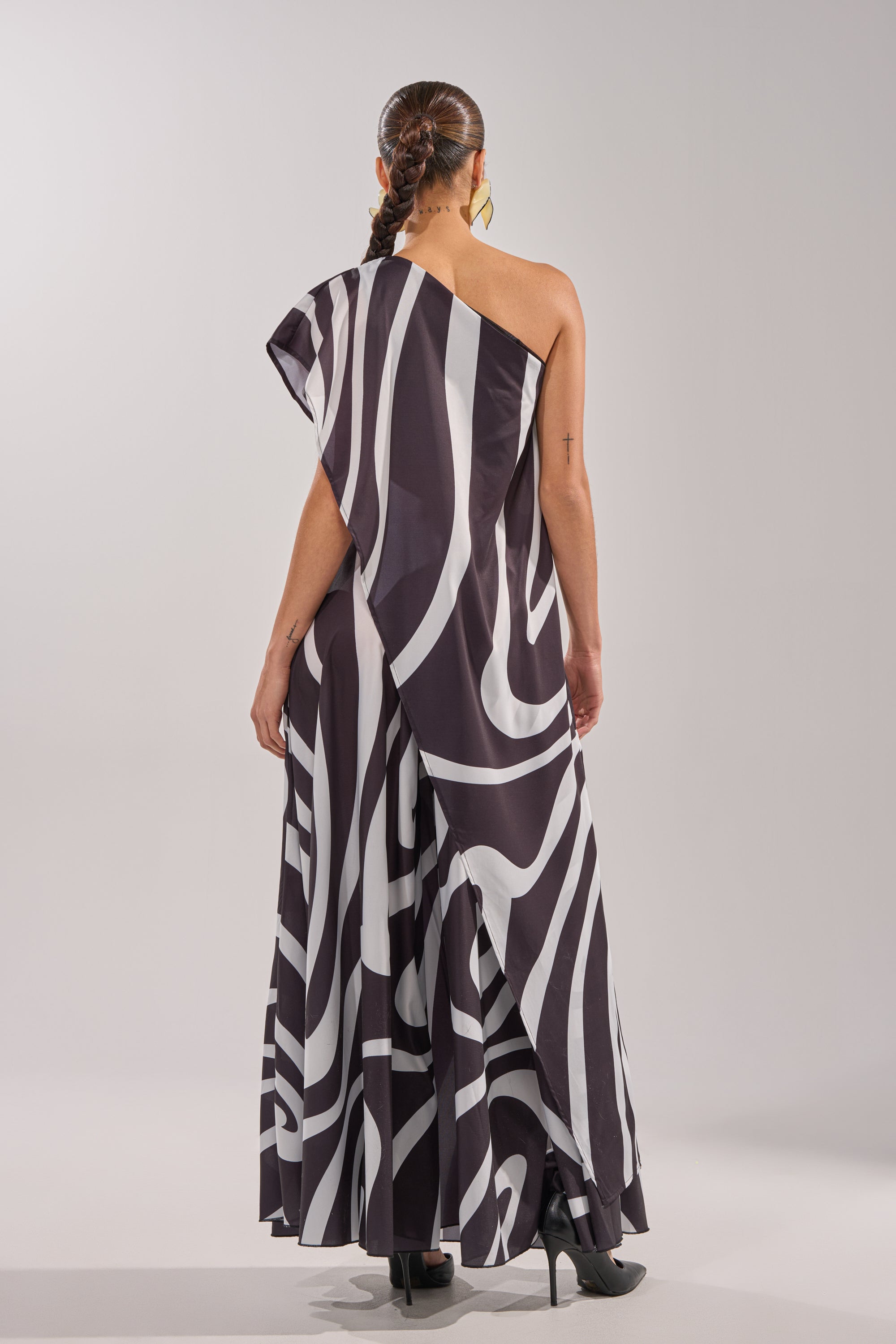A woman faces away, wearing the TRINA ELONGATED SINGLE SHOULDER TOP with a black and white abstract pattern, asymmetrical hem, black heels, hoop earrings, and two braids against a plain light gray background. 5