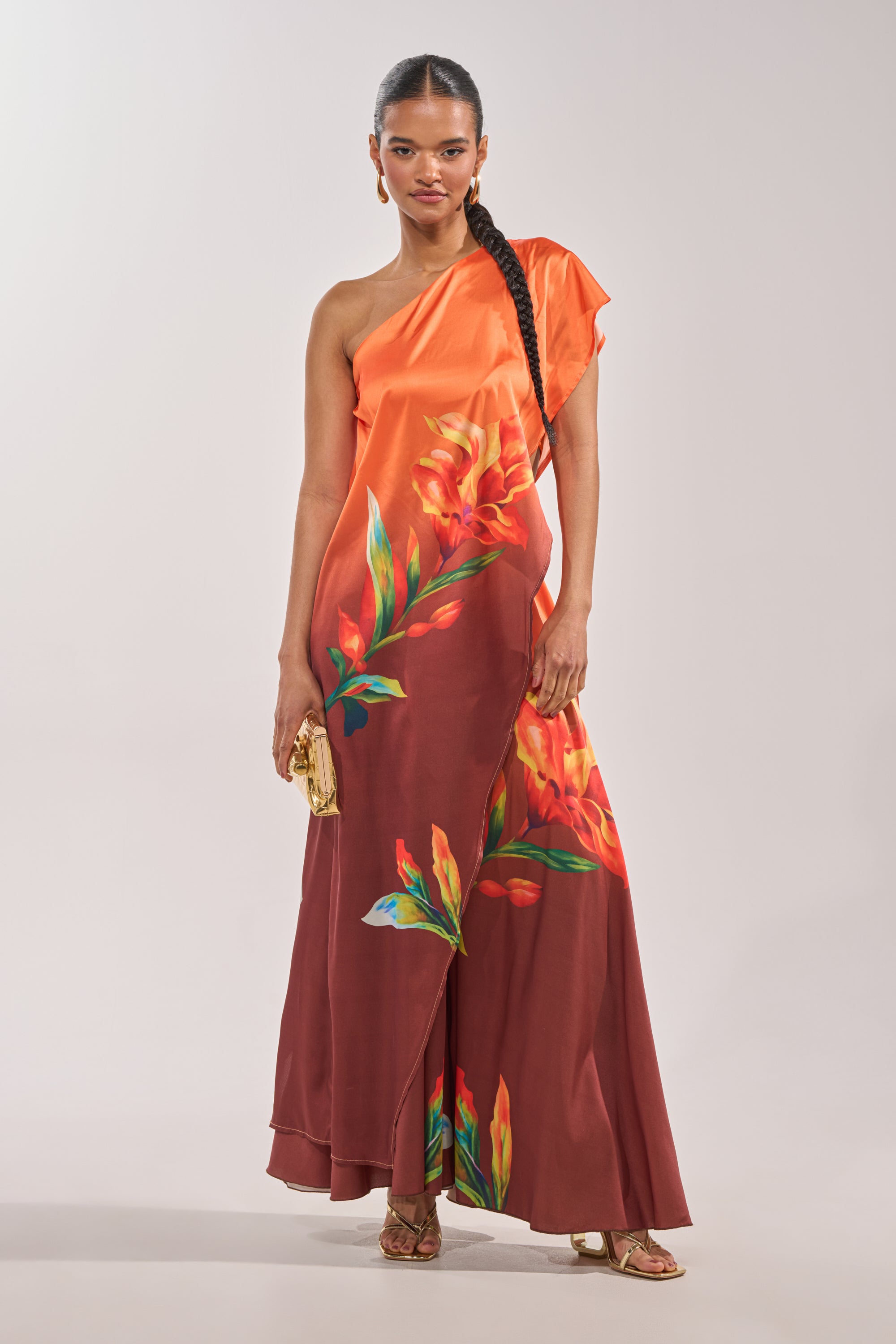 A woman in the SUNSET LOVER BLOUSE, styled with gold sandals, a gold clutch, and long braids, stands against a plain light background. 0