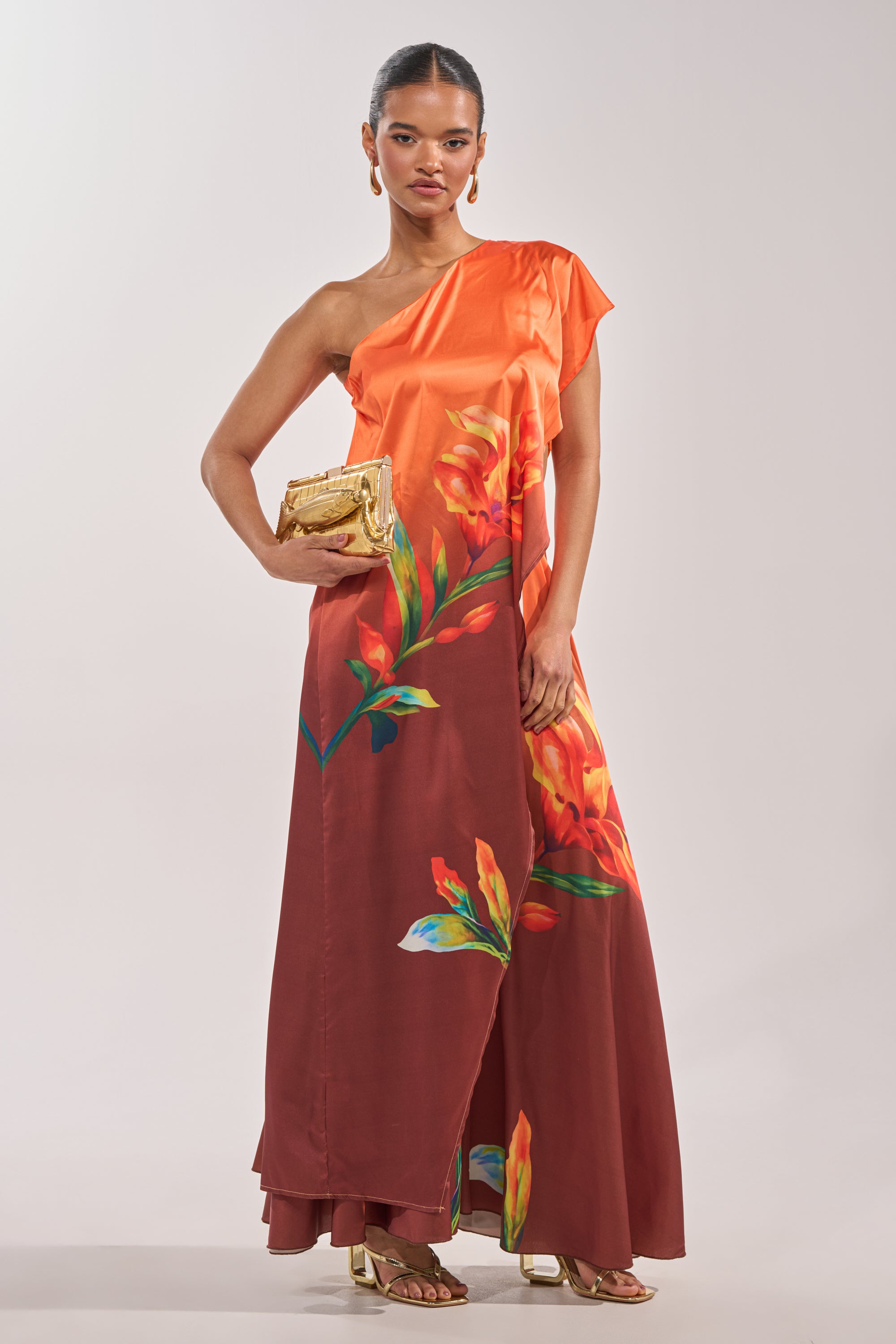 A woman poses wearing the SUNSET LOVER BLOUSE—an orange one-shoulder top paired with a maroon, floor-length skirt featuring bold floral prints. She accessorizes with a gold clutch, gold heels, and earrings. 1