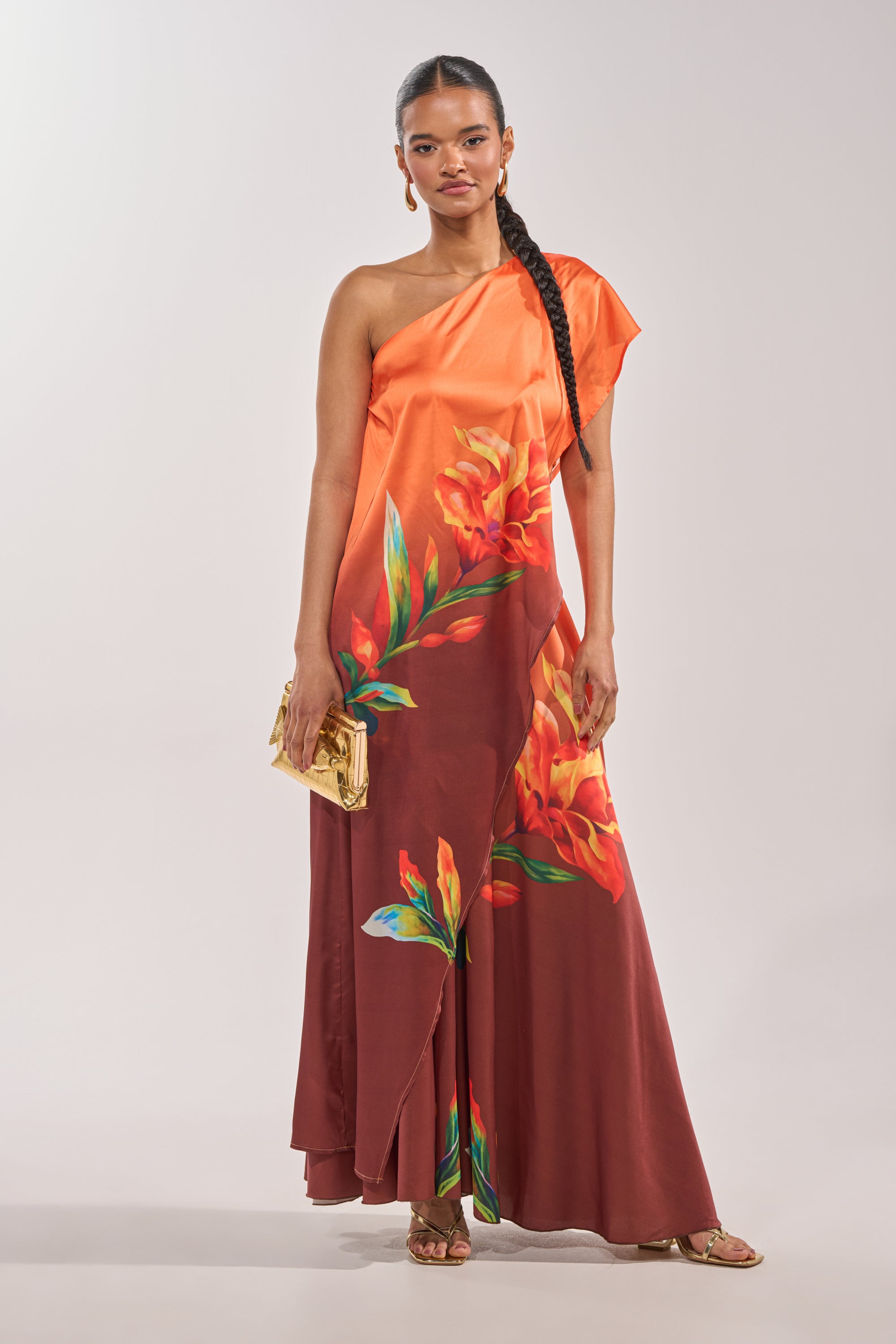 A woman wears the SUNSET LOVER BLOUSE—an orange, one-shoulder top paired with a maroon, floral-print floor-length skirt. She accessorizes with a gold clutch, gold sandals, and a sleek ponytail. 2