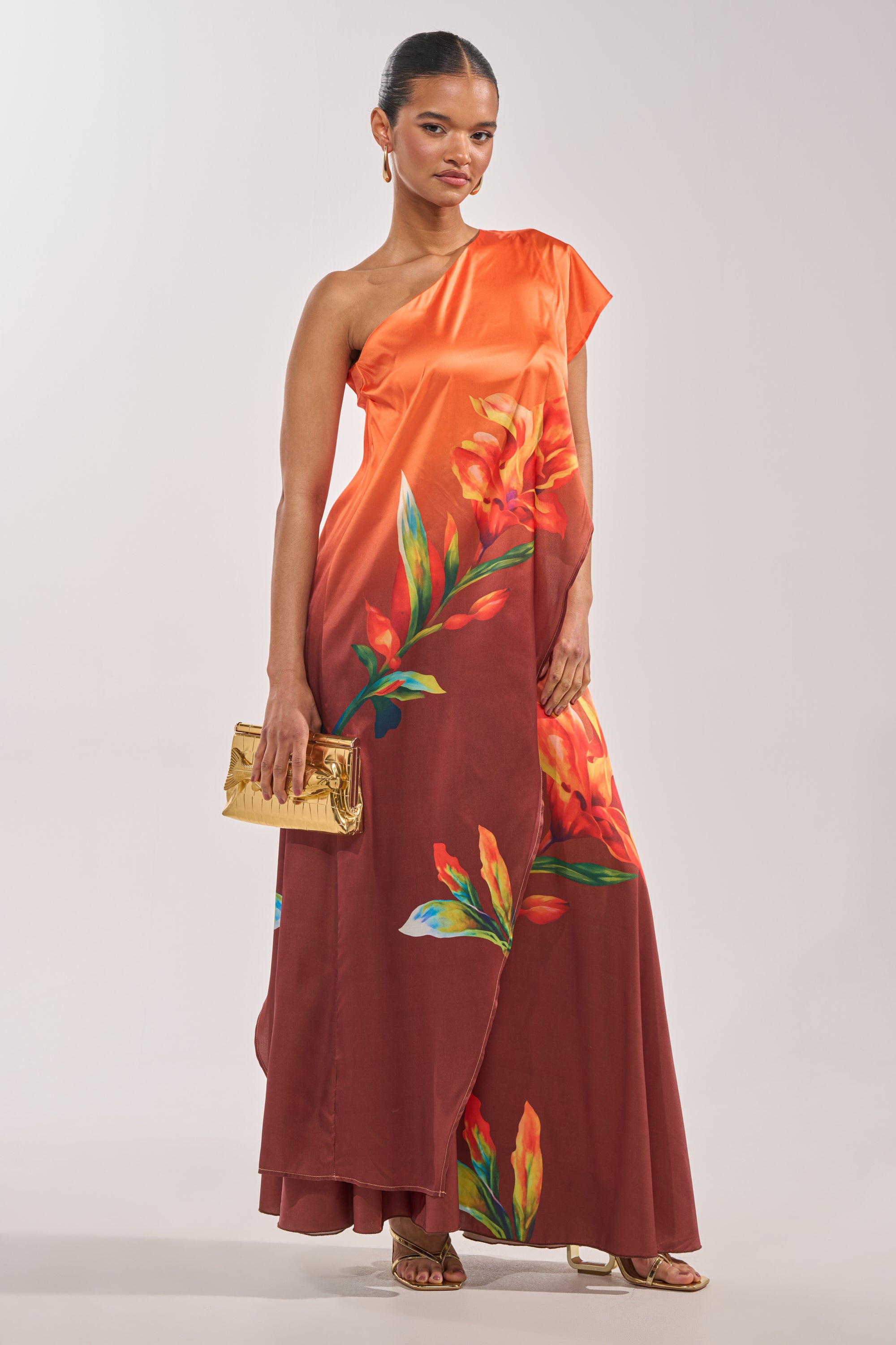 A woman in the SUNSET LOVER BLOUSE, with an orange to deep red gradient and tropical floral prints, holds a gold clutch. She accessorizes with slicked-back hair, gold earrings, and gold heels against a plain light background. 3