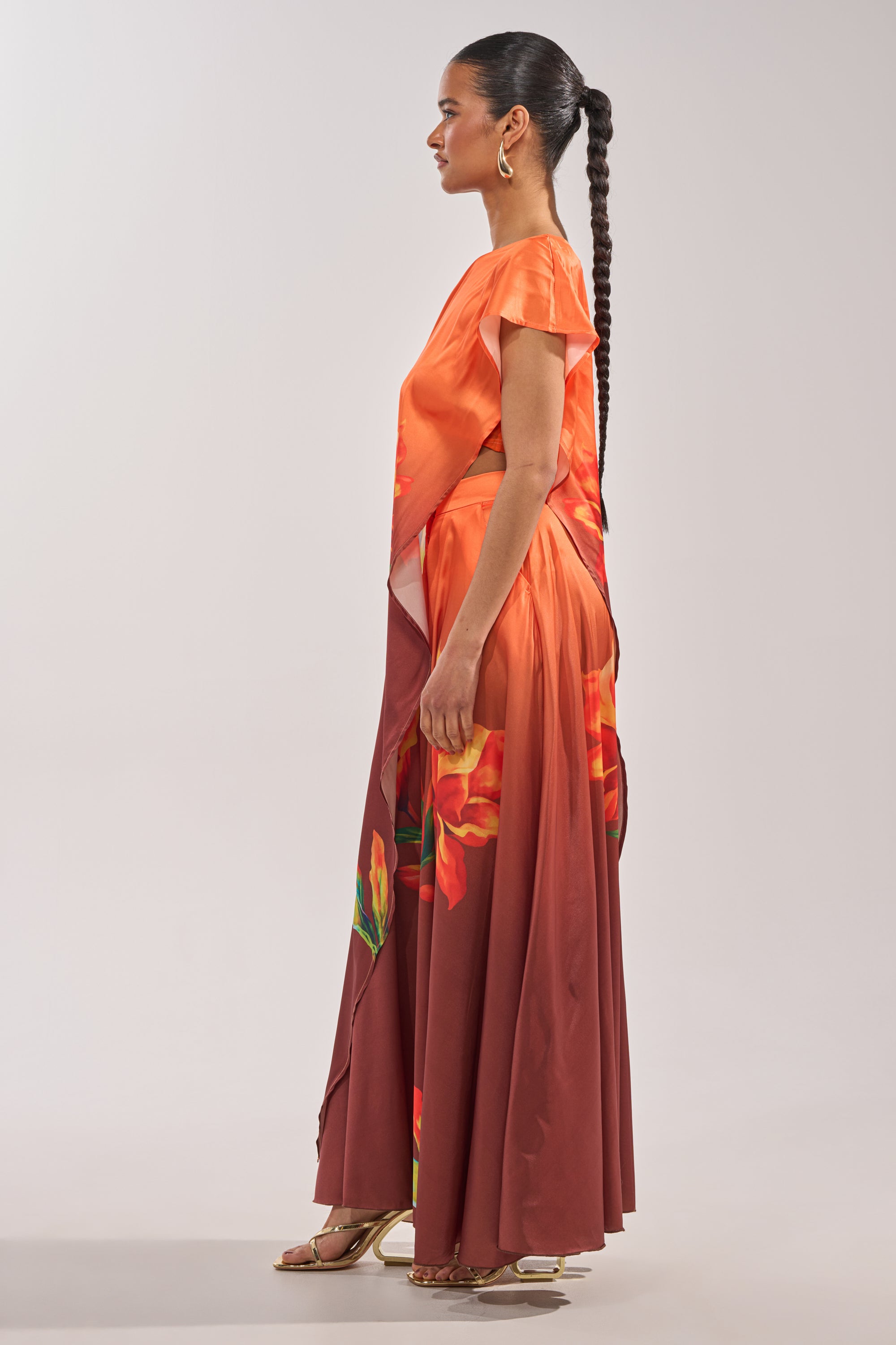 A woman stands in profile wearing the SUNSET LOVER BLOUSE, a flowing one-shoulder top in orange and brown with large floral prints. She pairs it with gold sandals and a long braided ponytail against a plain, light background. 4