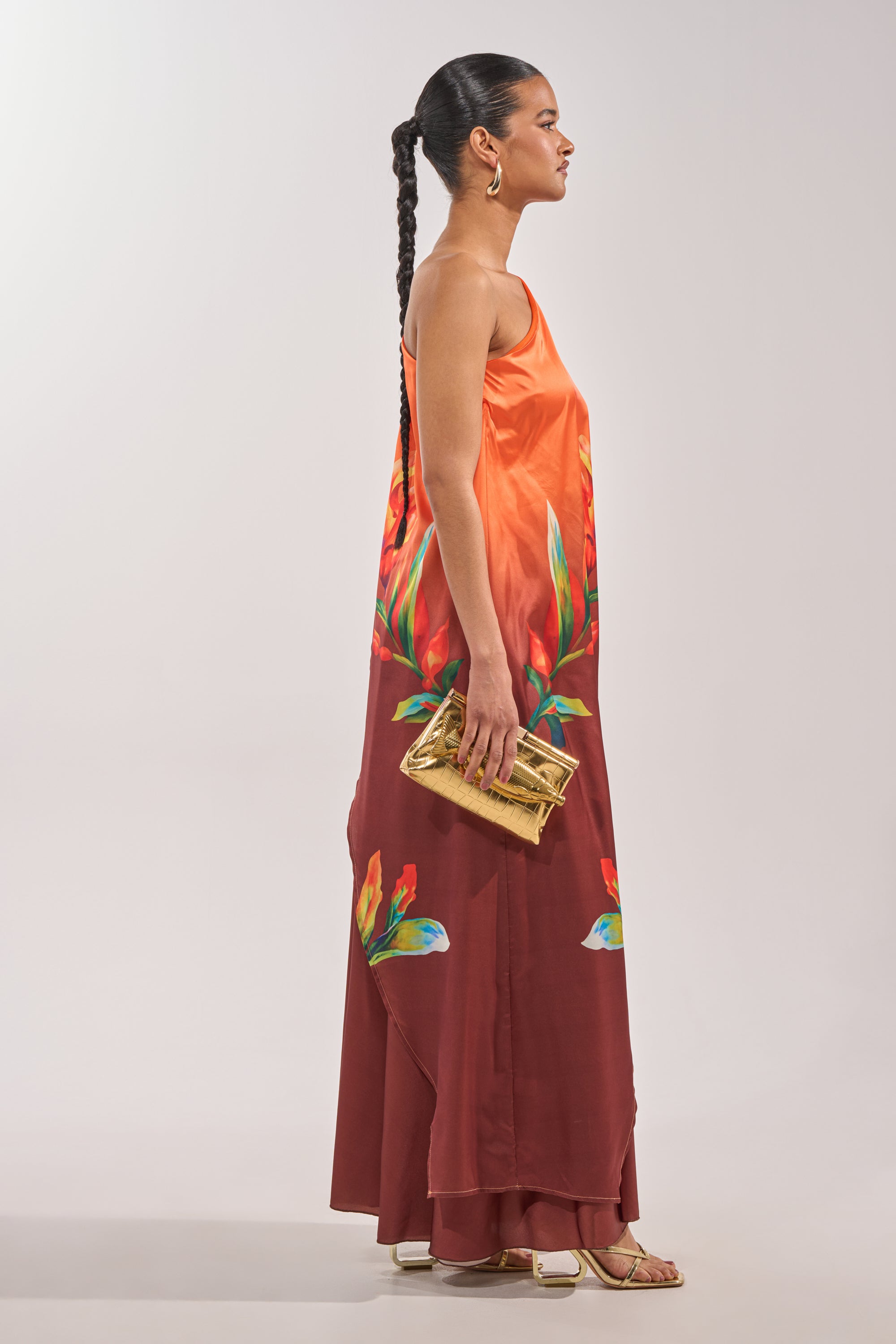 A woman with a long braid stands in profile, wearing a sleeveless orange and maroon floral maxi dress styled as elegantly as the SUNSET LOVER BLOUSE, paired with hoop earrings, white sandals, and a gold clutch against a plain background. 5