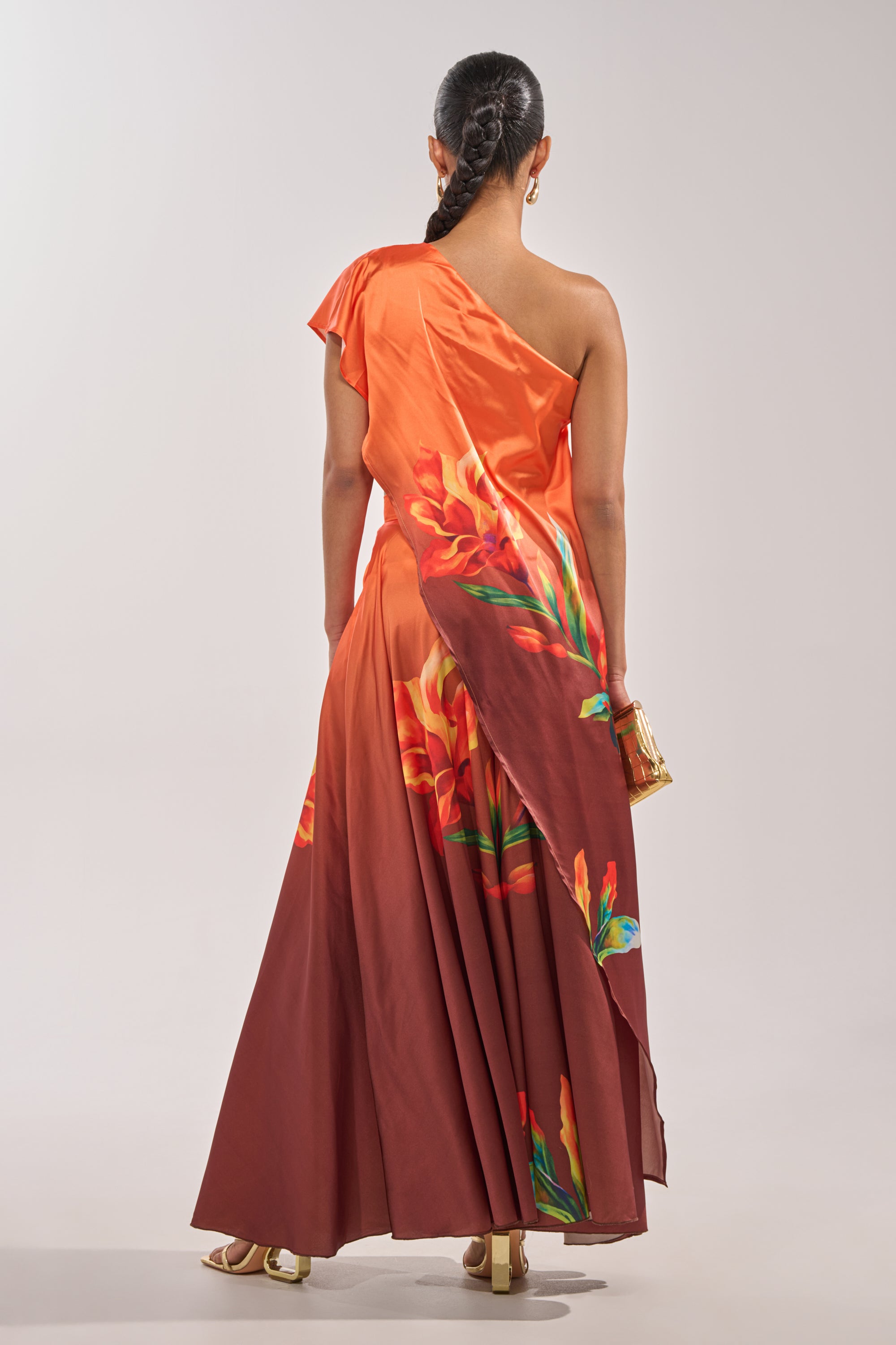 A woman with a braided ponytail stands facing away, wearing the SUNSET LOVER BLOUSE in orange and maroon floral print, paired with gold heels and holding a small gold clutch. 6
