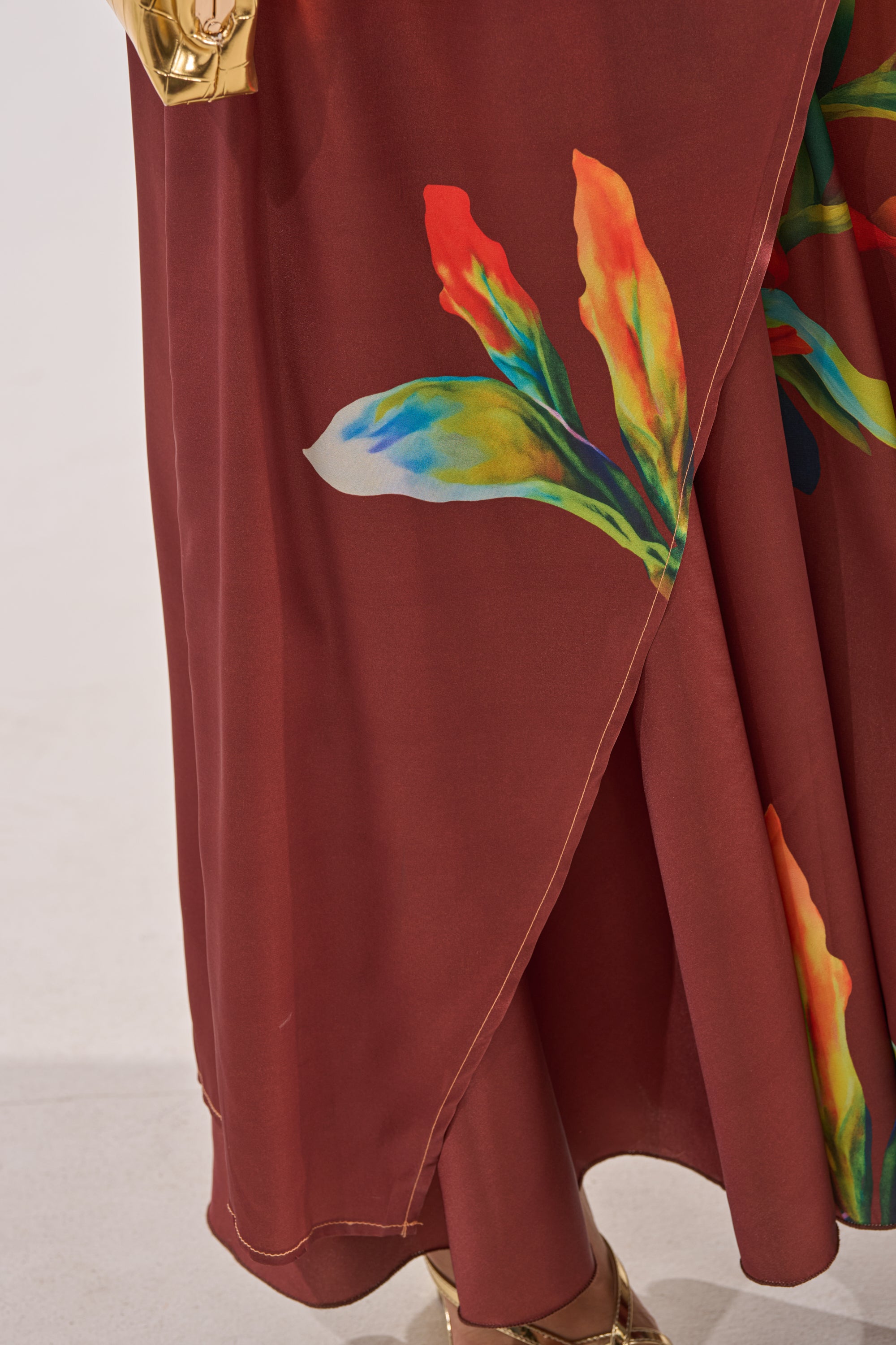 A close-up of a maroon skirt with a vibrant floral print, paired stylishly with the asymmetrical satin SUNSET LOVER BLOUSE, plus a gold bracelet and gold shoes partially visible. 8