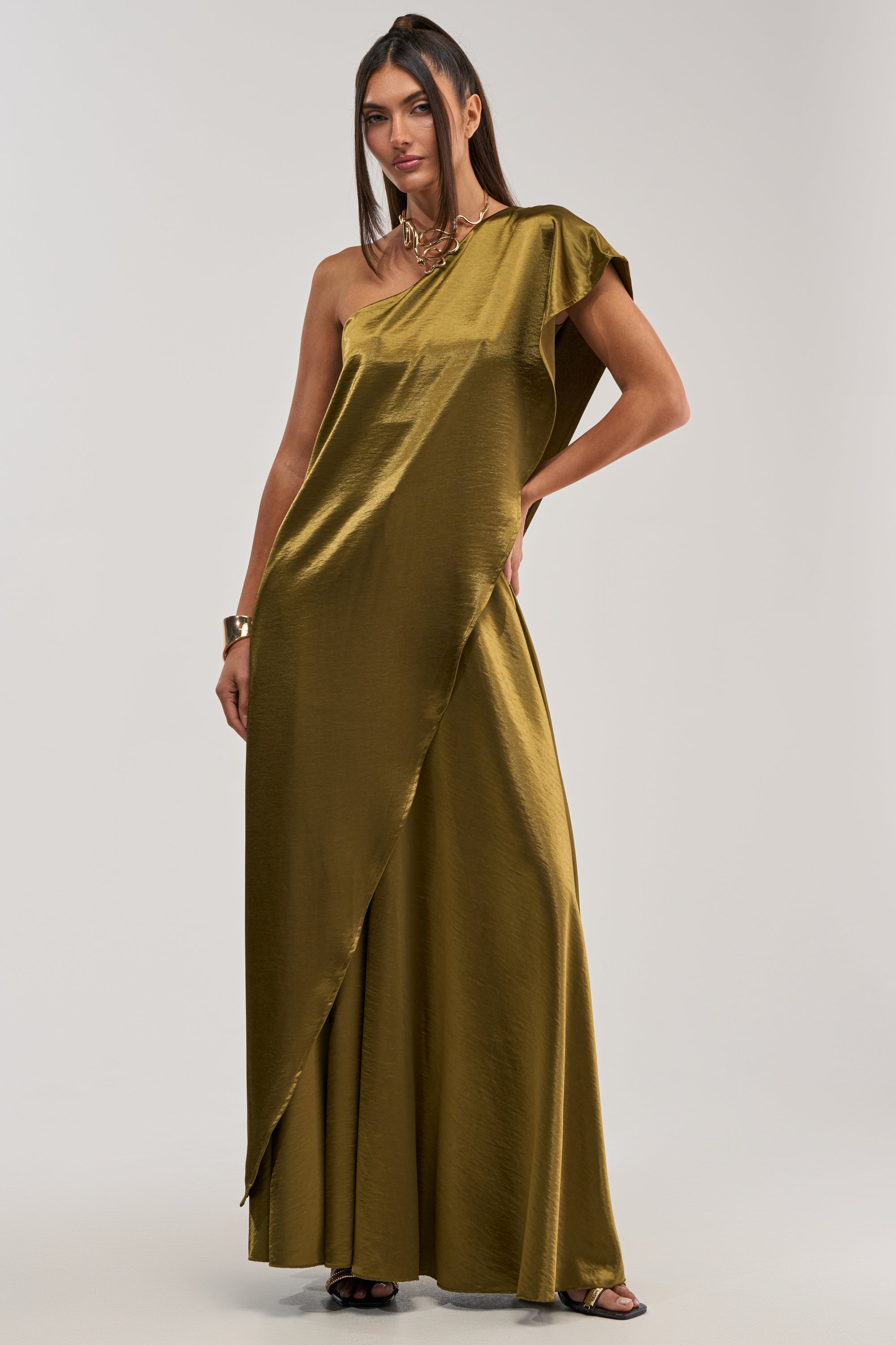 A woman models the TRUE GODDESS ONE-SHOULDER TOP in shiny olive satin, styled as a floor-length gown with an asymmetrical sleeve. She accessorizes with black heels, a chunky necklace, and bracelet against a plain light background.