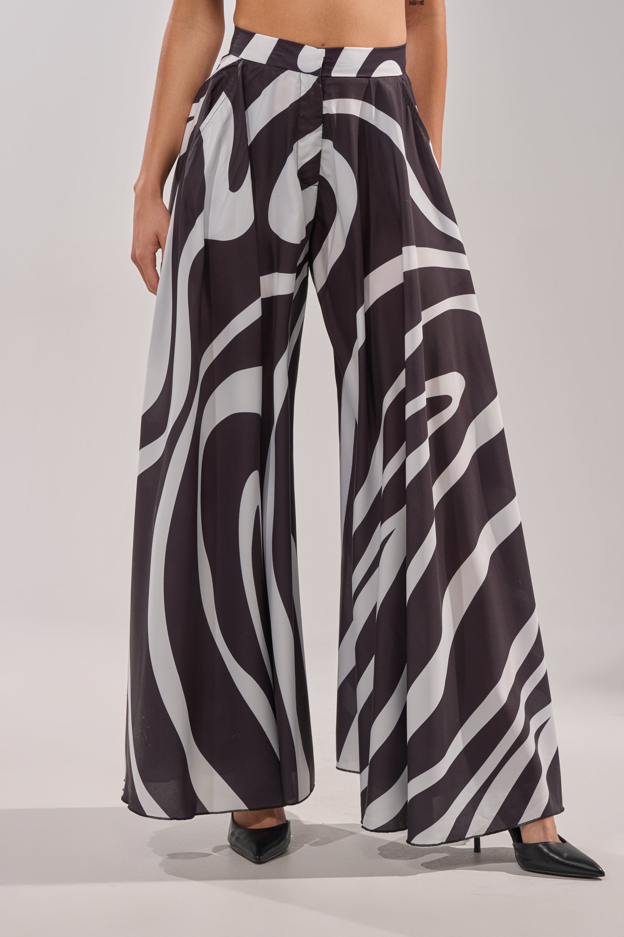 Someone is wearing TRINA PLEATED PALAZZO PANT featuring high-rise swirl print with bold white abstract patterns, paired with black pointed-toe heels, standing against a neutral background. 0