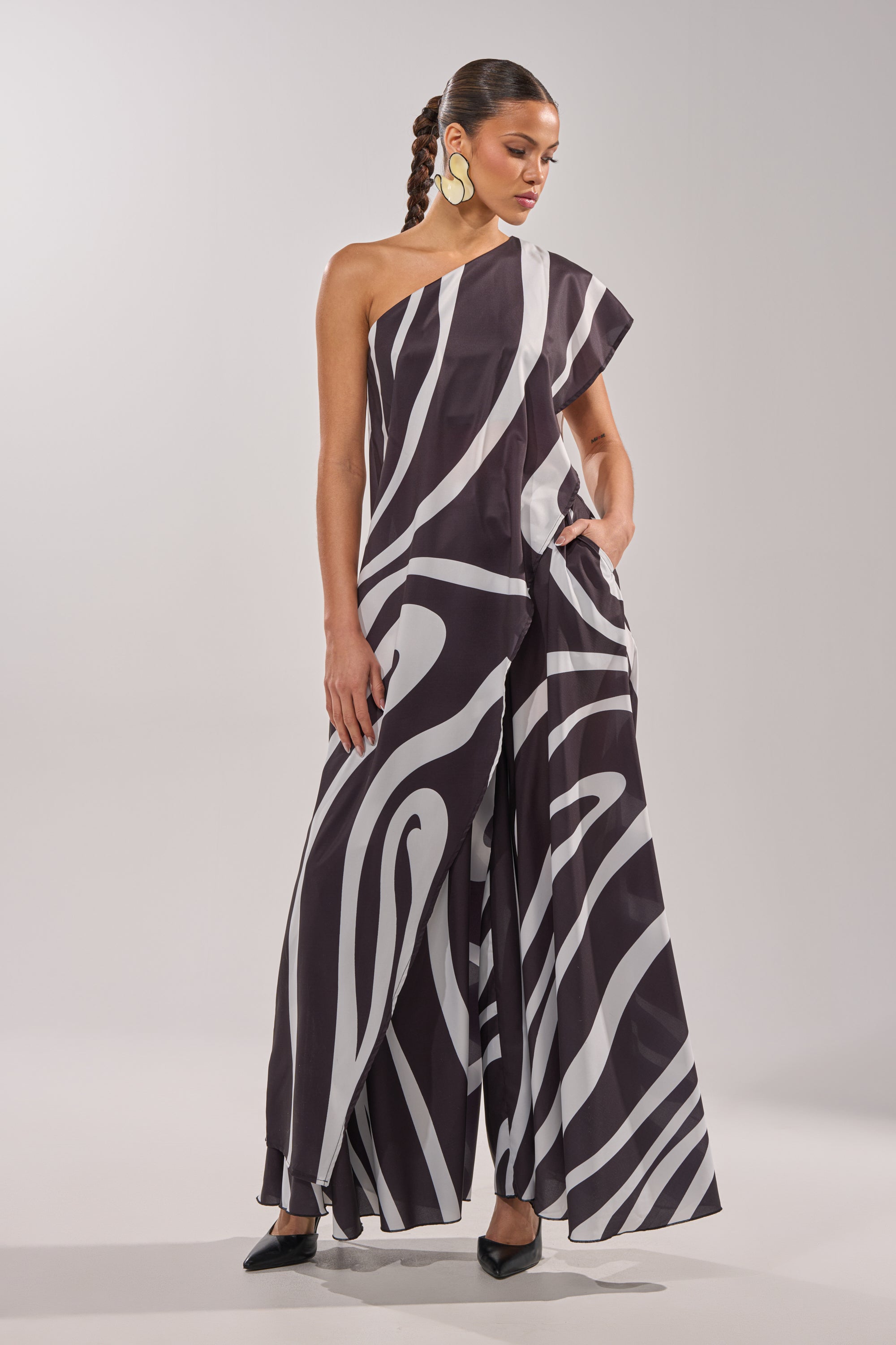 A woman poses confidently against a plain background, wearing the TRINA PLEATED PALAZZO PANT—a high-rise, wide-leg style with a bold black and white swirl print—styled with braided hair, large gold earrings, and black pointed heels. 8
