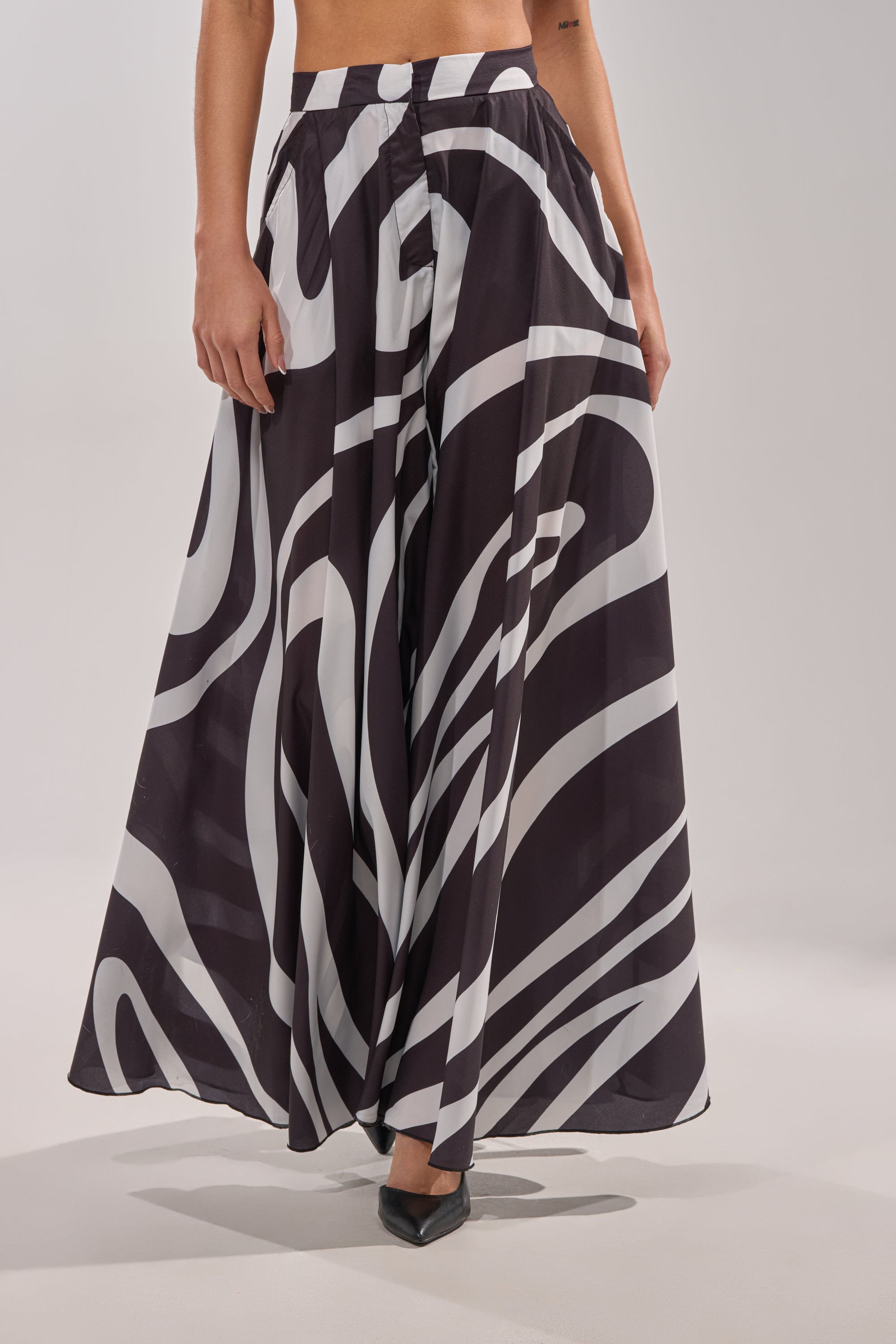 A person wears the TRINA PLEATED PALAZZO PANT, featuring a high-rise waist and bold black and white swirl print, styled with pointed black heels against a neutral background. 1