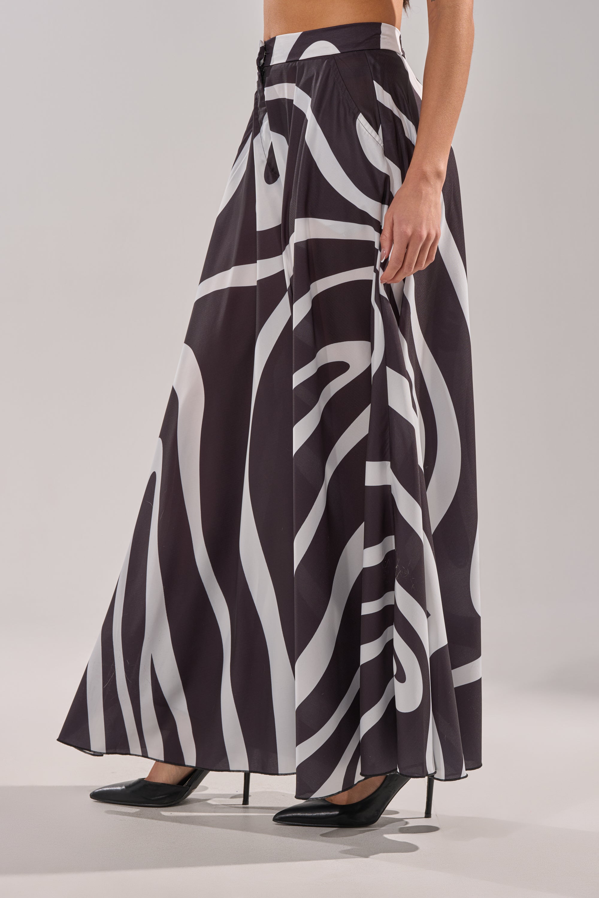 A woman wearing the TRINA PLEATED PALAZZO PANT in a black and white swirl print with black high heels, standing against a light gray background. 2