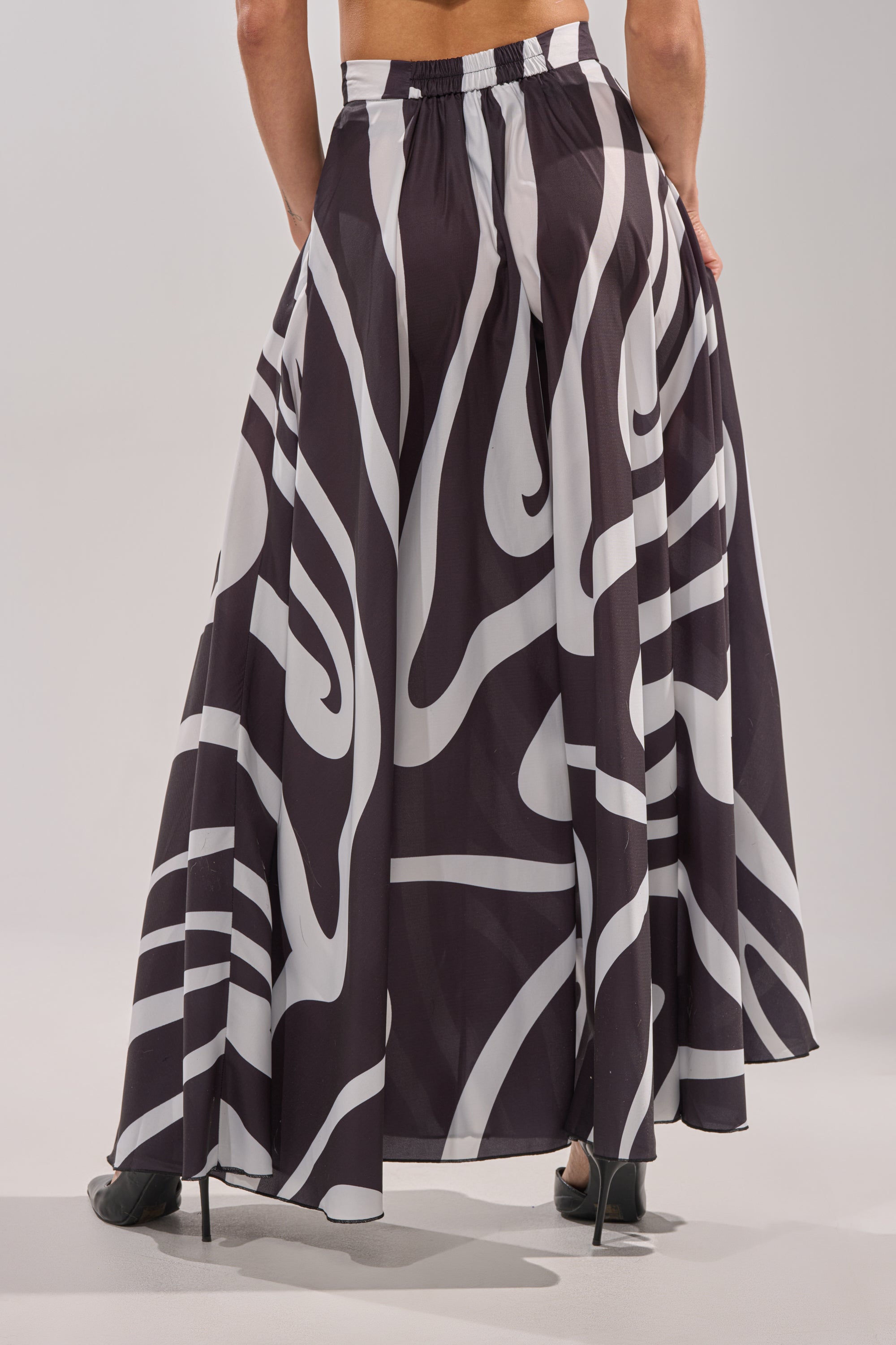 A person facing away showcases the TRINA PLEATED PALAZZO PANT, featuring a high-rise waist and black-and-white swirl print; only their lower back and black high-heeled shoes are visible against a plain background. 3