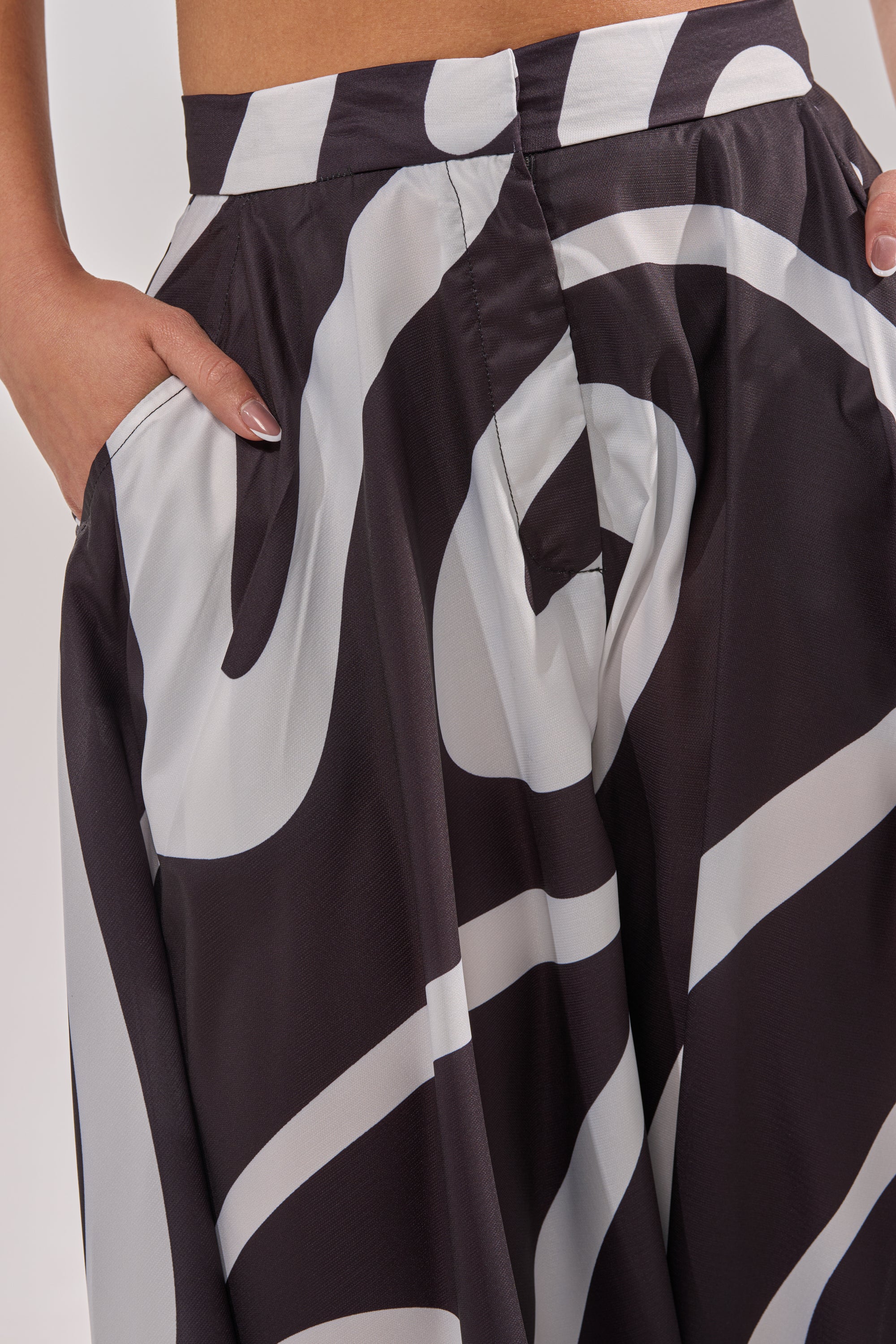 Someone wearing the TRINA PLEATED PALAZZO PANT, a high-rise, wide-leg style with a bold black and white swirl print, hands in pockets, shown from waist to mid-thigh. 6
