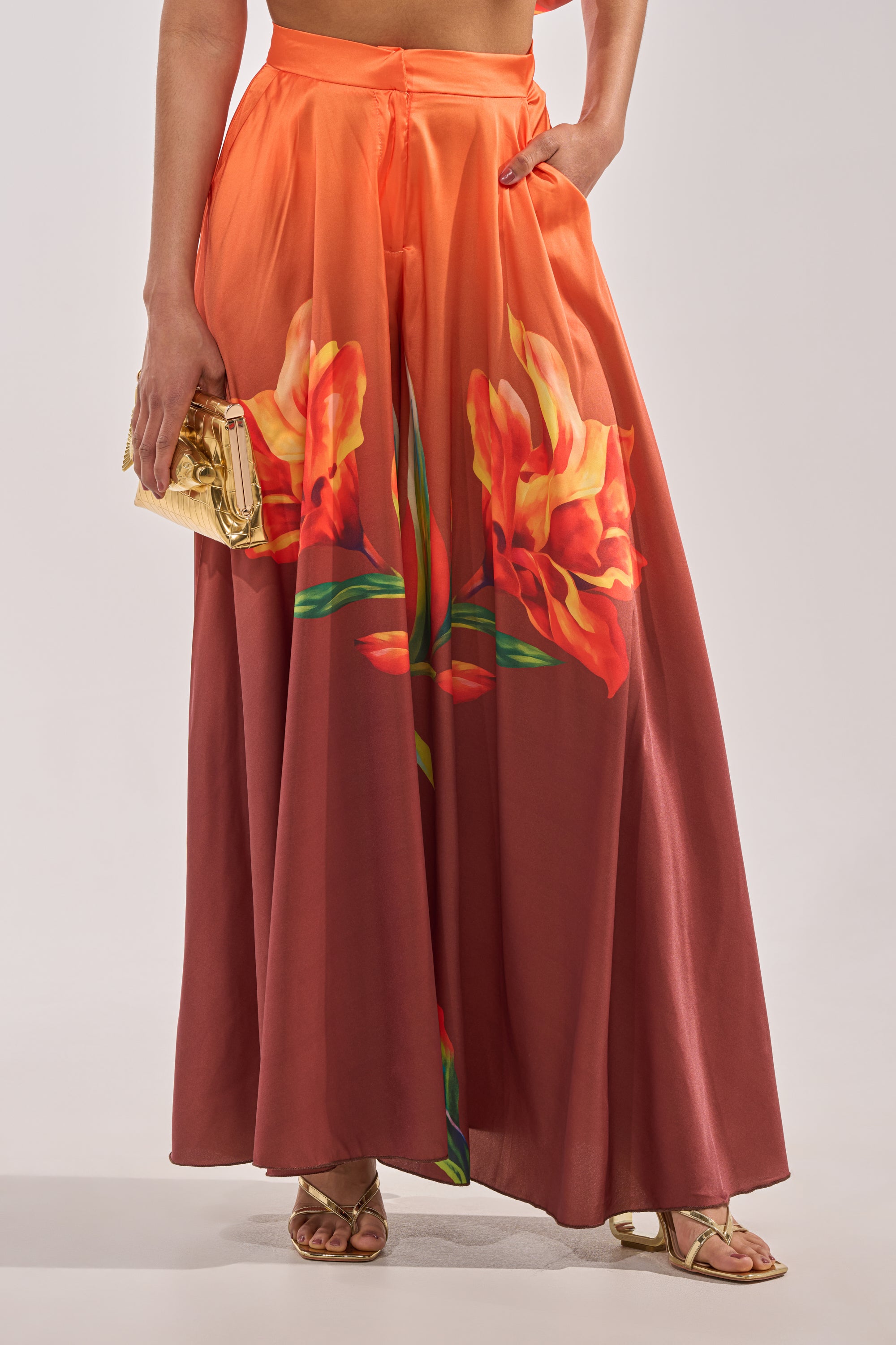 Someone wearing the SUNSET LOVER PALAZZO PANT, featuring a bold orange and yellow tropical floral print, paired with gold sandals and a small gold clutch. Their hand rests in the pocket of the pants. 0