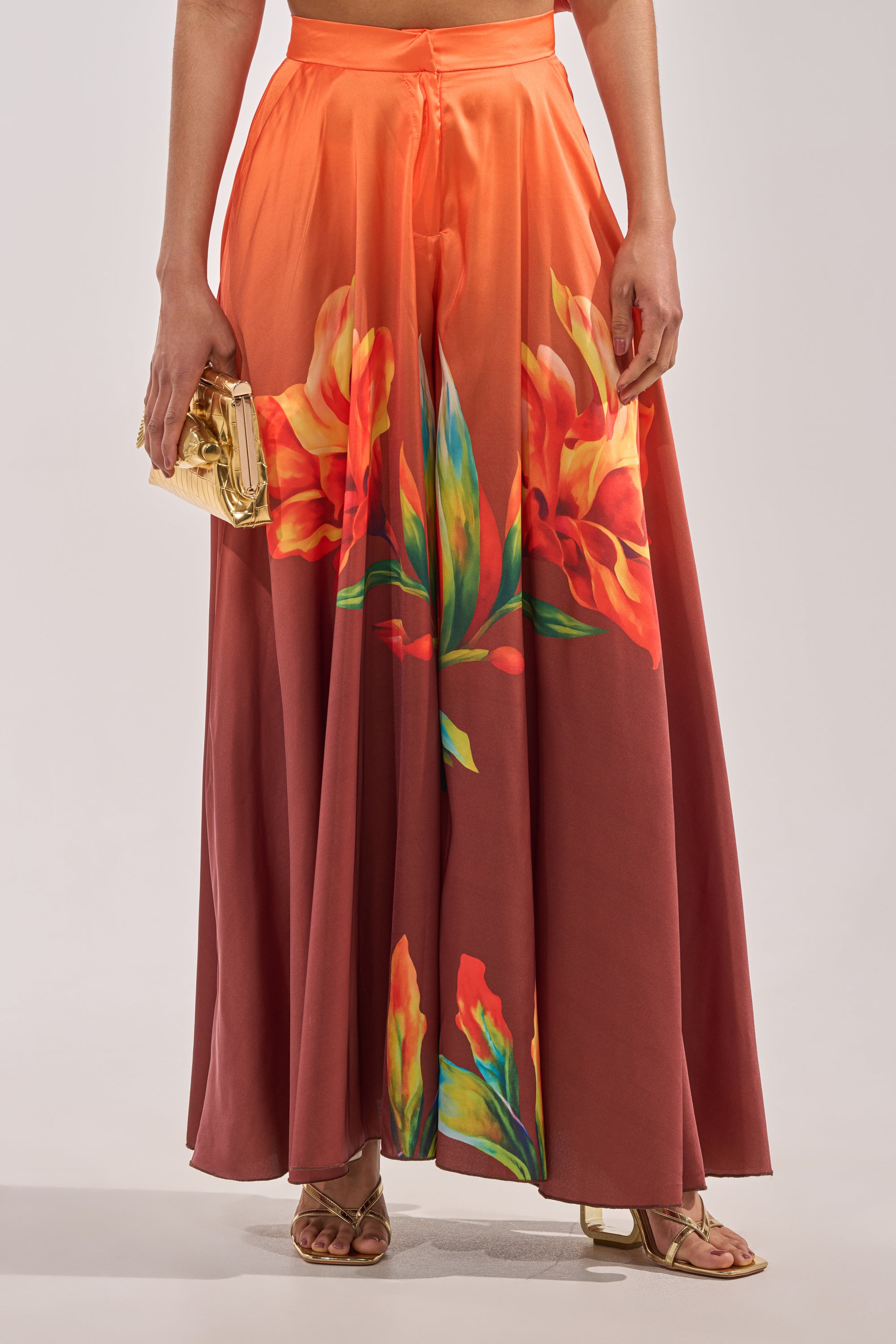 Someone wearing the SUNSET LOVER PALAZZO PANT—flowing, high-waisted satin pants with bold floral prints—paired with a small gold clutch and gold sandals. 1