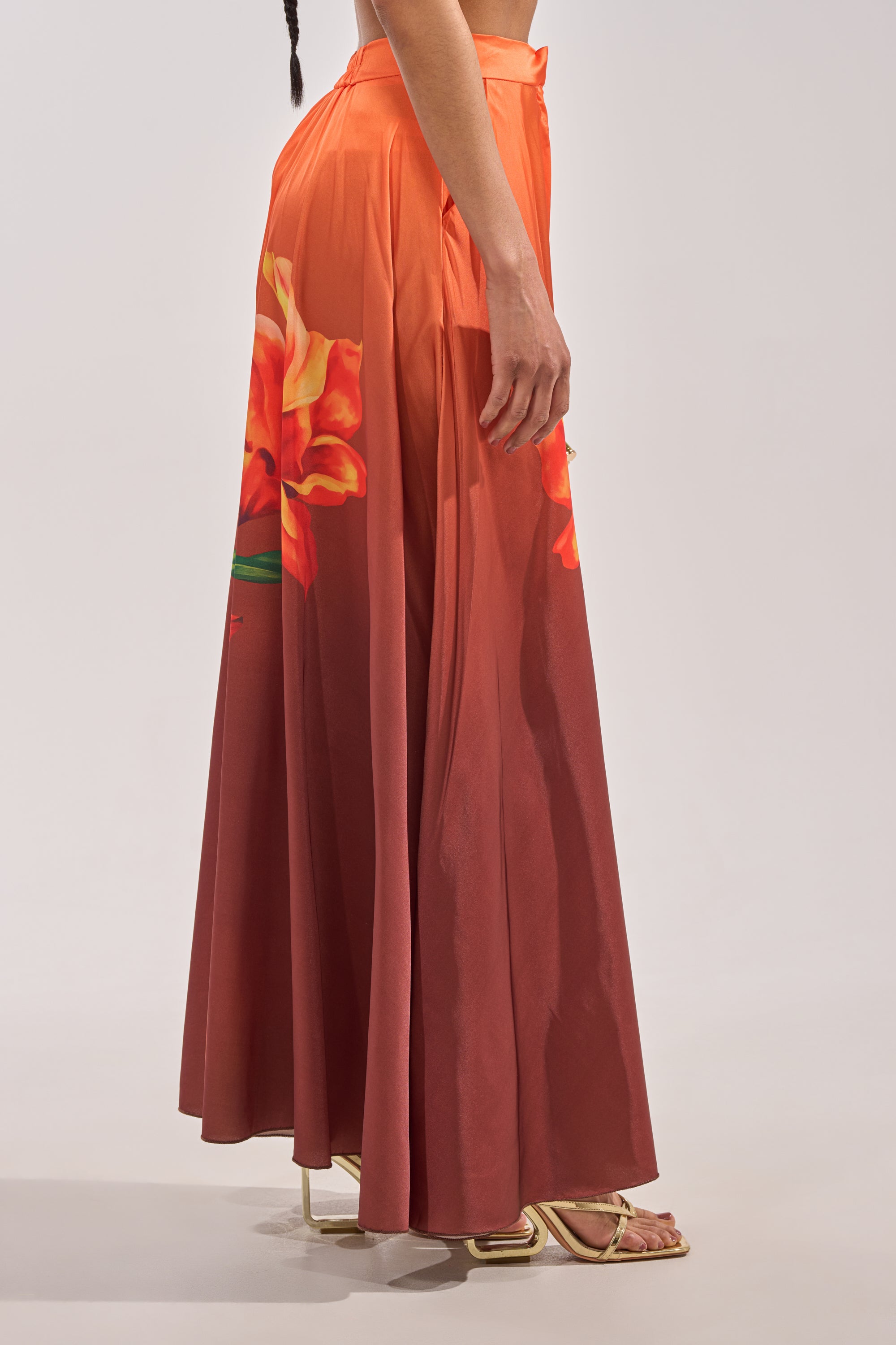 A person in profile wears the SUNSET LOVER PALAZZO PANT, a long, flowy style with bold tropical prints in reddish-orange, paired with strappy heeled sandals for an effortless, breezy look. 2