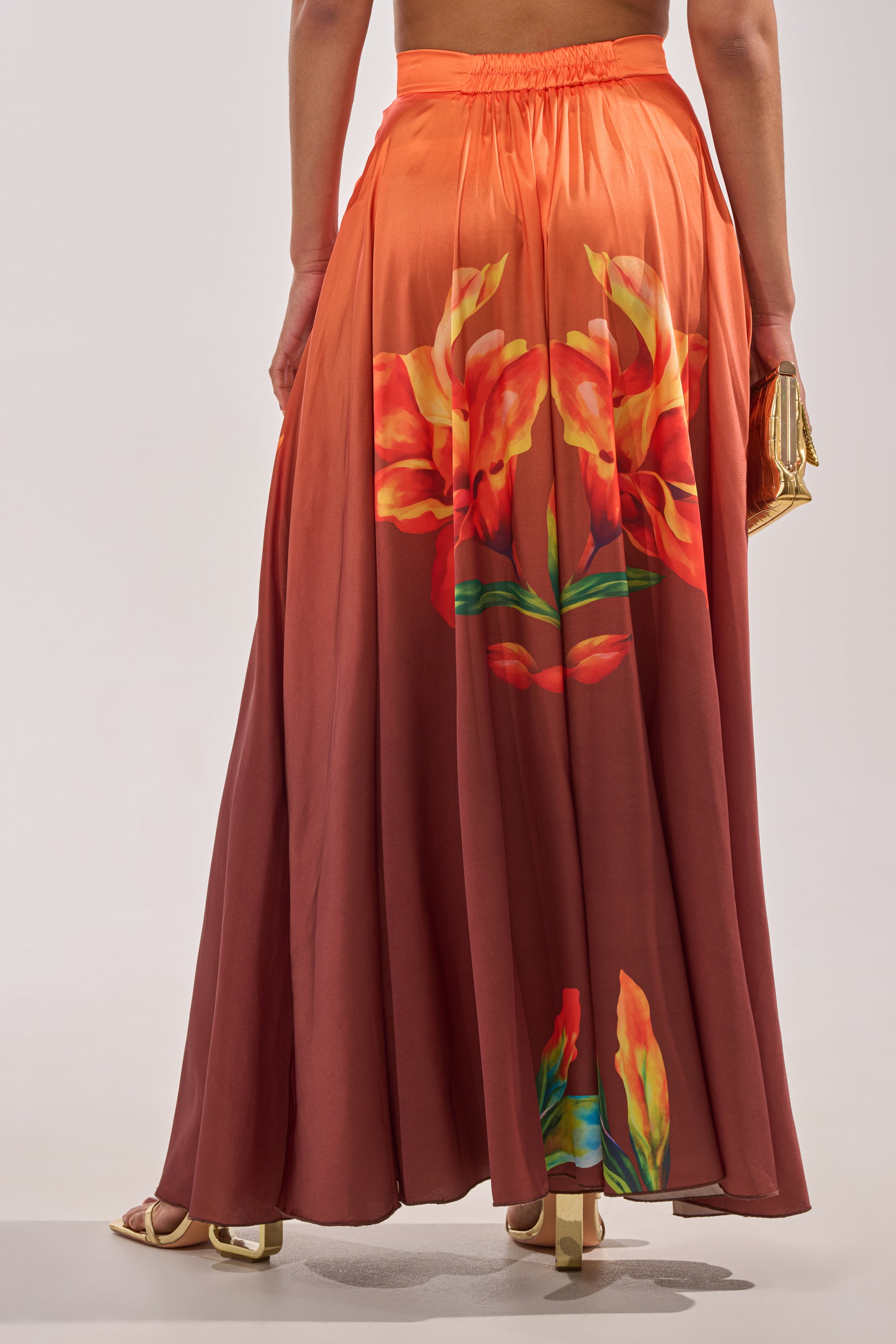 A person models the SUNSET LOVER PALAZZO PANT—wide-leg, orange-red pants with bold floral back prints—styled with heeled sandals and a gold clutch. The upper body is mostly out of frame. 3