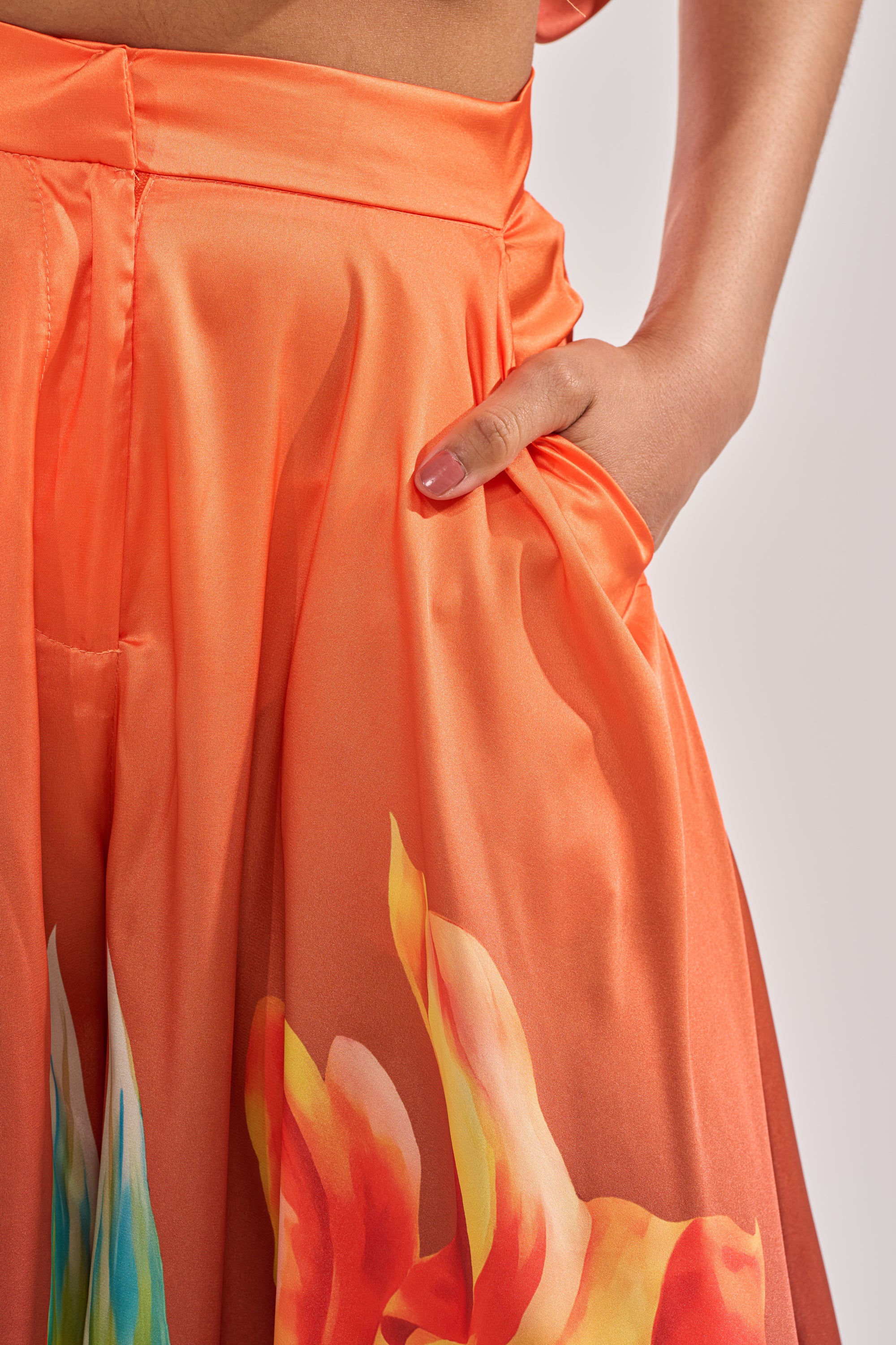 A person models the SUNSET LOVER PALAZZO PANT, featuring vibrant floral print and orange satin fabric. The shot highlights the waist and upper legs, emphasizing the pant’s sheen and bold pattern, with one hand tucked in a pocket. 6