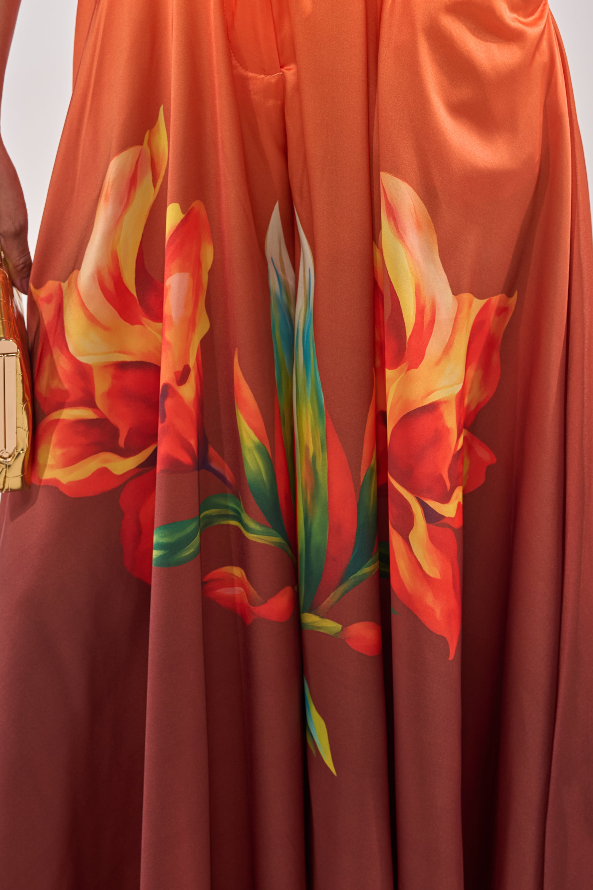 A close-up shows the SUNSET LOVER PALAZZO PANT in shiny satin peeking out on the left, next to a gold rectangular purse, alongside a flowing reddish-orange dress with a bold floral print in yellow, green, and red. 7