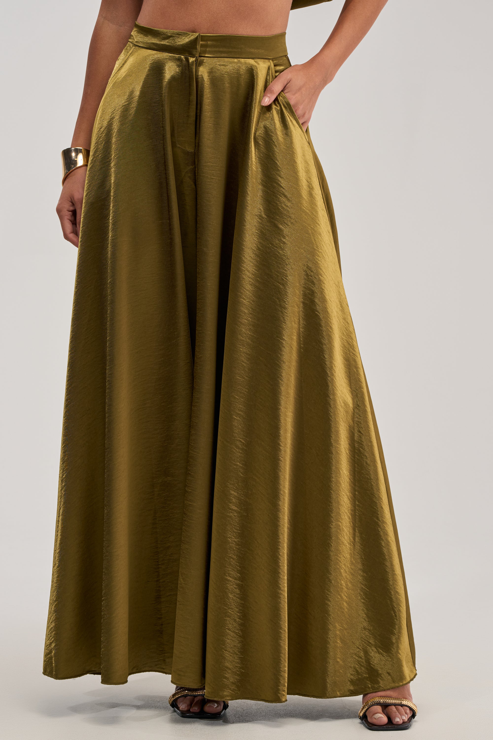 A person wears the TRUE GODDESS GREEN PALAZZO PANT in olive satin, featuring a wide-leg silhouette, high-rise waist, and side pockets, styled with a gold cuff bracelet and open-toe heels. The upper body is mostly out of frame.