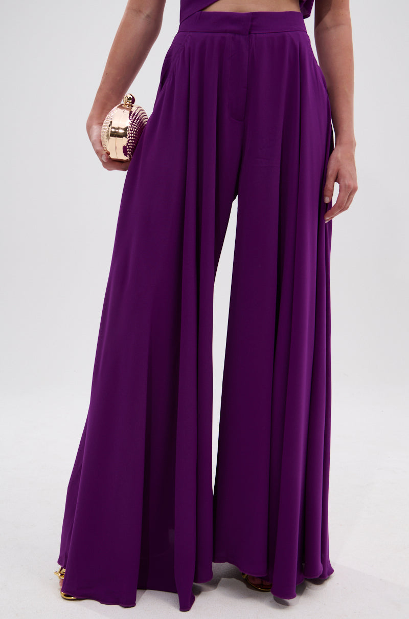 A person wearing the TRINA PLEATED PALAZZO PANT in vibrant violet and holding a metallic clutch stands against a plain light background. 1
