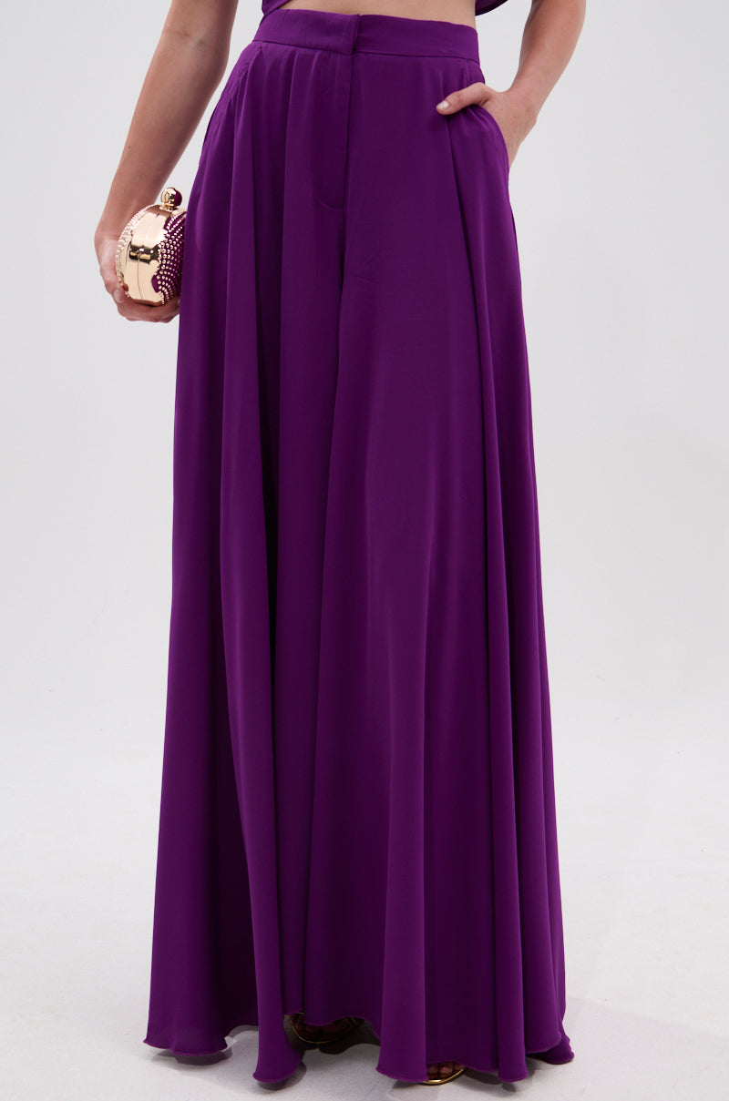 Wearing the TRINA PLEATED PALAZZO PANT in vibrant violet, a person poses with a gold clutch and one hand in their pocket, midriff partially visible against a light background. 2