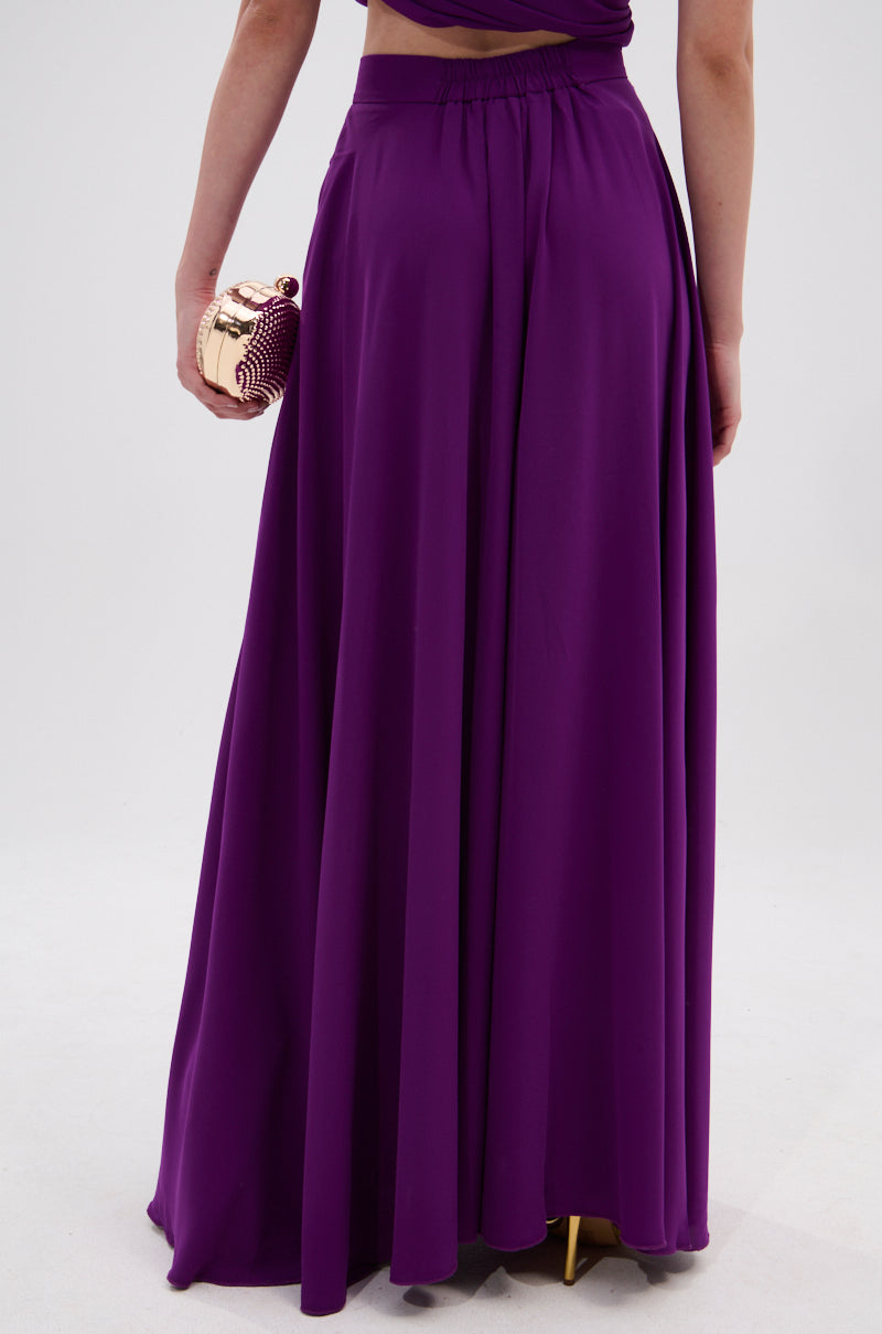 A person wearing high-rise, flowing violet TRINA PLEATED PALAZZO PANT stands with their back to the camera, holding a small shiny clutch bag. The background is plain and light-colored. 4