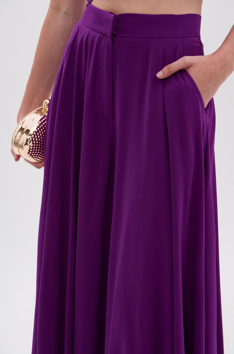 A person models the TRINA PLEATED PALAZZO PANT in vibrant violet, one hand in their pocket, holding a small round gold and burgundy beaded purse against a plain background. 8