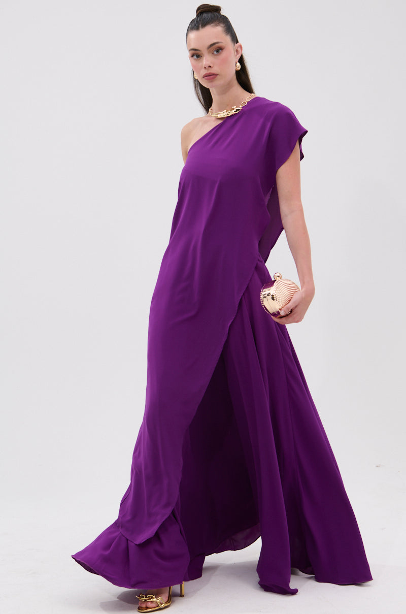 Wearing the TRINA ELONGATED SINGLE SHOULDER TOP in purple, a woman with sleek dark hair and a chunky gold necklace pairs it with gold heels and a round gold clutch, posing against a plain white background. 1