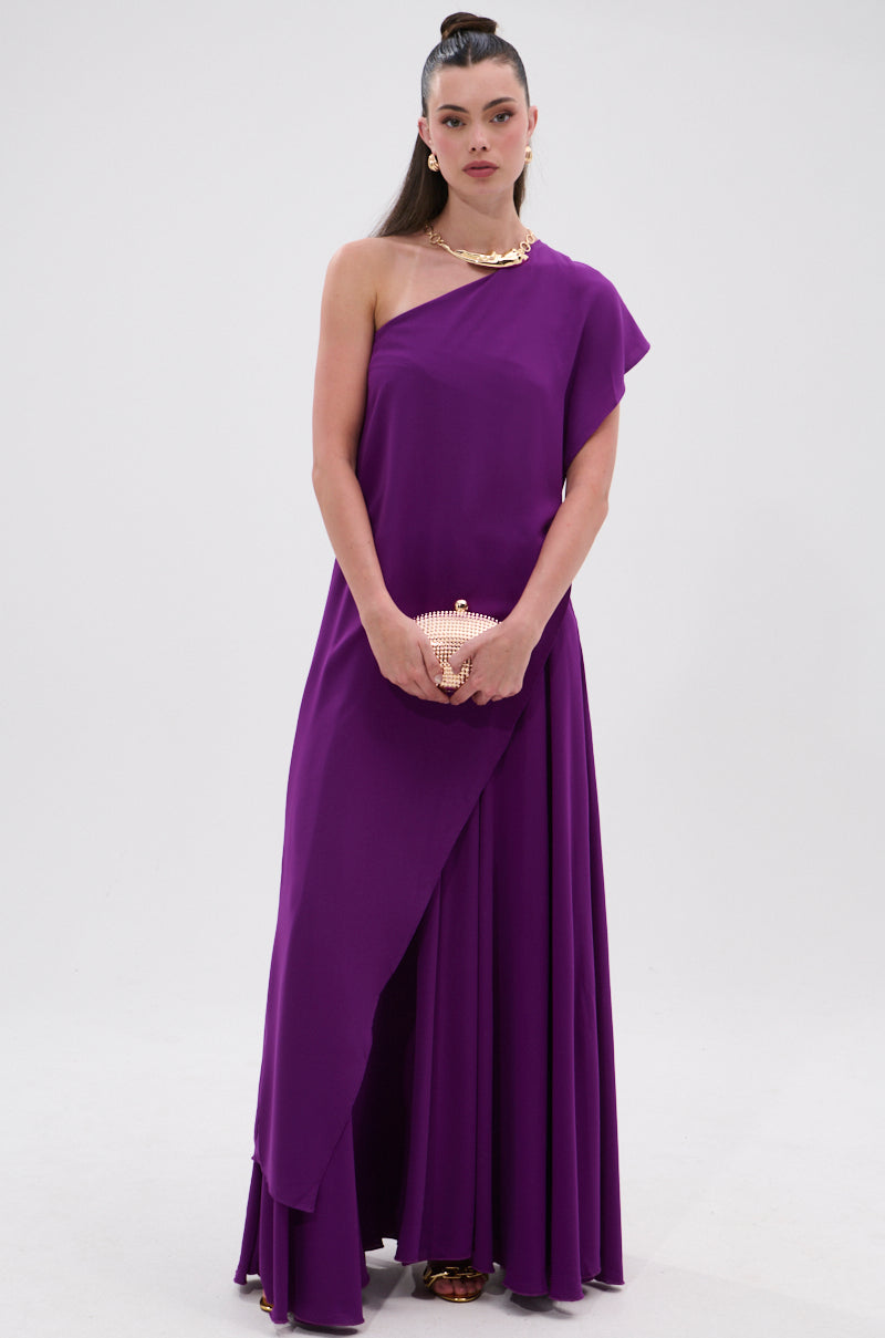 A woman stands against a white background wearing the TRINA ELONGATED SINGLE SHOULDER TOP in purple. She holds a gold clutch, wears a chunky gold necklace, and has her hair styled in a sleek ponytail. 2