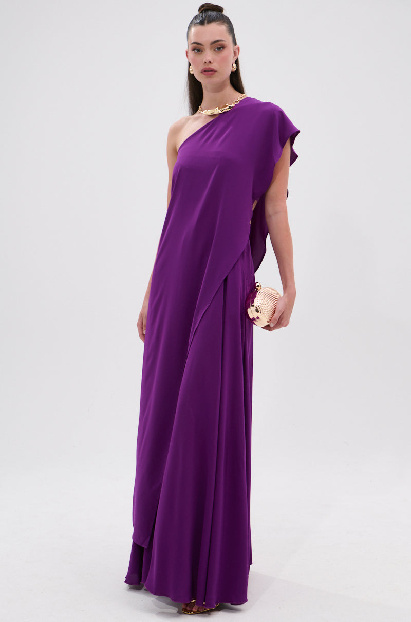 A woman stands against a white background wearing the TRINA ELONGATED SINGLE SHOULDER TOP with a floor-length purple skirt, gold necklace, and matching gold clutch. 3