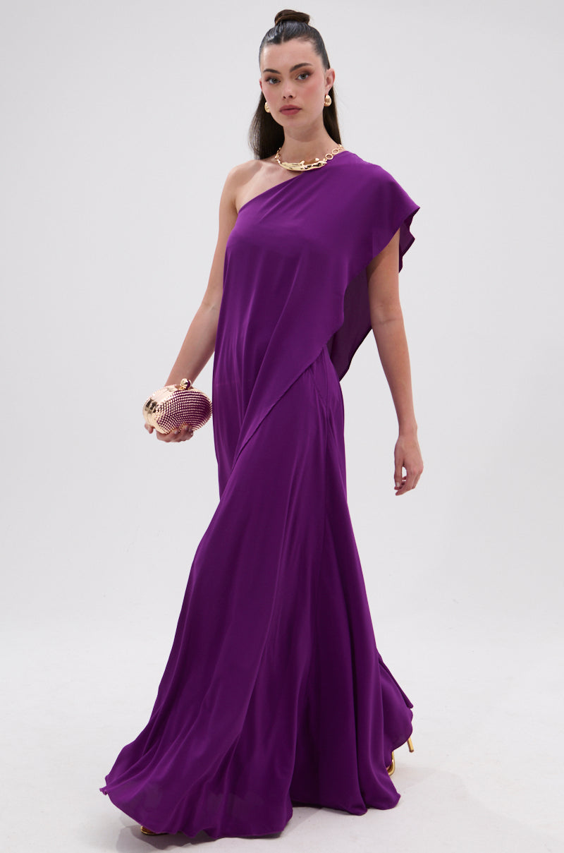 Wearing the TRINA ELONGATED SINGLE SHOULDER TOP in purple, paired with a textured gold clutch and a chunky gold necklace, a person stands against a plain light background. 4