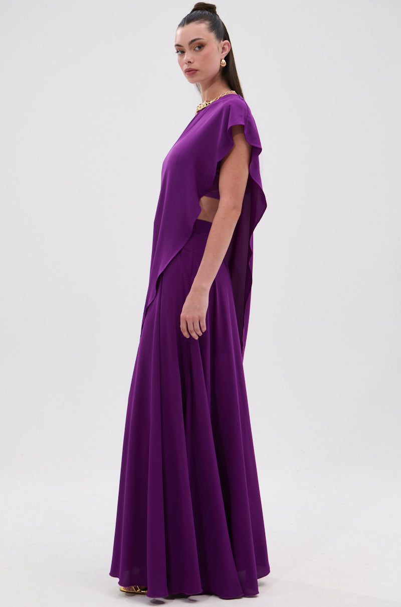 A woman stands in profile against a white background, wearing the TRINA Elongated Single Shoulder Top in purple with an asymmetrical one-shoulder silhouette and open side. Her long hair is pulled back, and she accessorizes with a gold necklace. 6