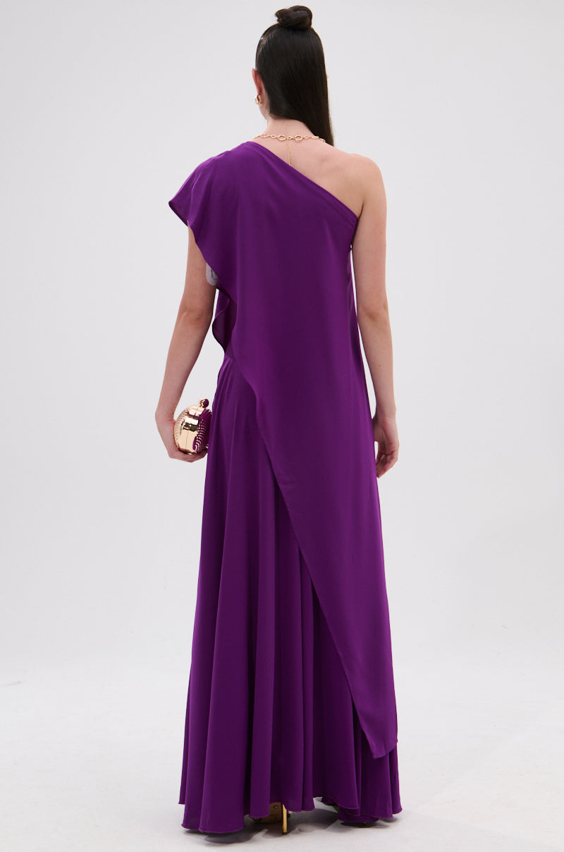 A woman faces away, showcasing the TRINA Elongated Single Shoulder Top in purple with an asymmetrical silhouette. She holds a gold clutch and wears her long dark hair in a topknot against a plain white background. 7