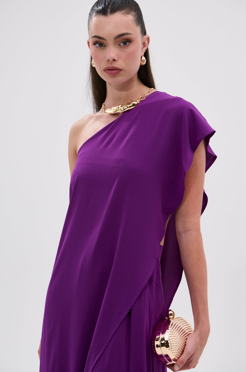 A woman with straight dark hair pulled back wears the TRINA ELONGATED SINGLE SHOULDER TOP in vibrant purple, paired with a chunky gold necklace and small gold clutch. She gazes confidently at the camera against a plain background. 8