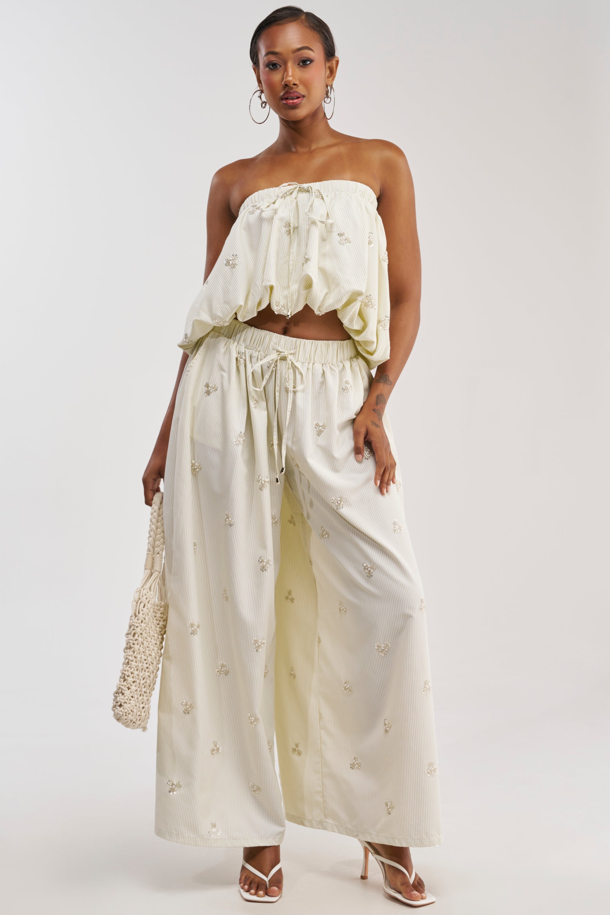 A woman stands against a plain background wearing the LIGHT AND AIRY POPLIN PANT—a pair of loose cream summer pants with small floral embroidery. She pairs them with a matching strapless top, white heels, and carries a woven bag.