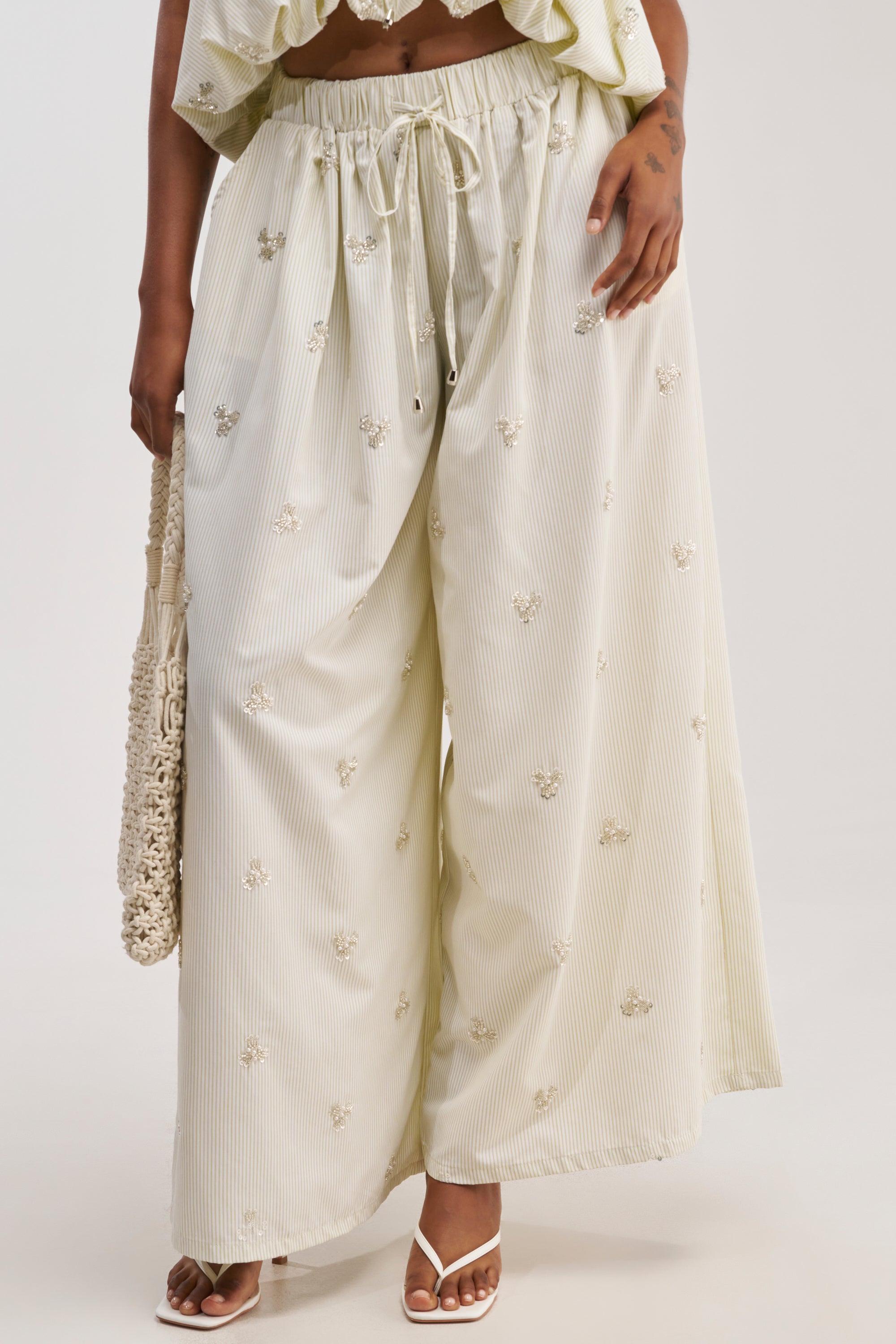 A person is shown from the waist down wearing the LIGHT AND AIRY POPLIN PANT, featuring beige stripes with subtle vertical patterns and delicate embroidered florals. They are holding a woven bag and wearing white heeled sandals.