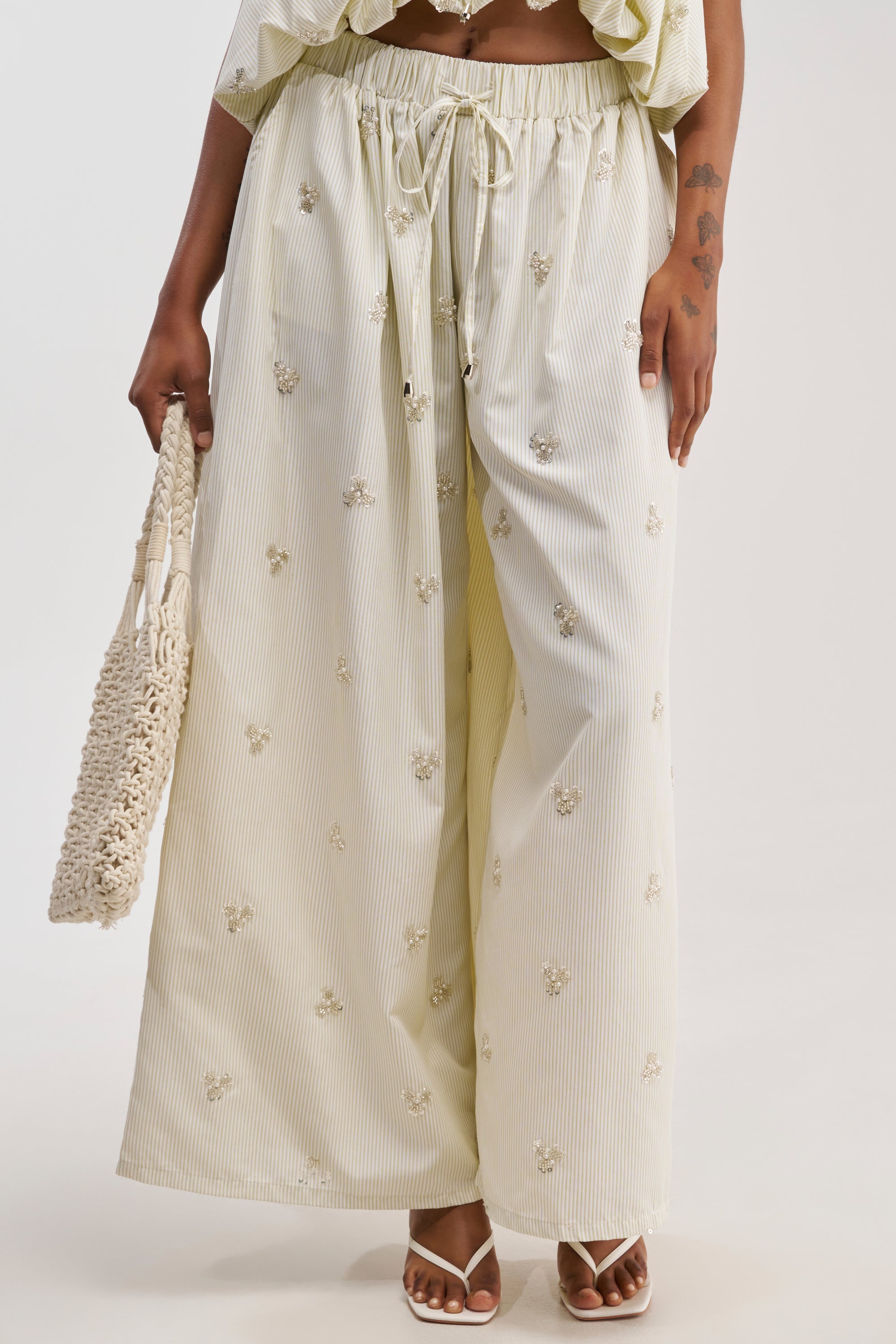 Wearing the LIGHT AND AIRY POPLIN PANT with embroidered details, the person—whose lower body is shown—carries a woven bag and pairs the look with open-toed heels.