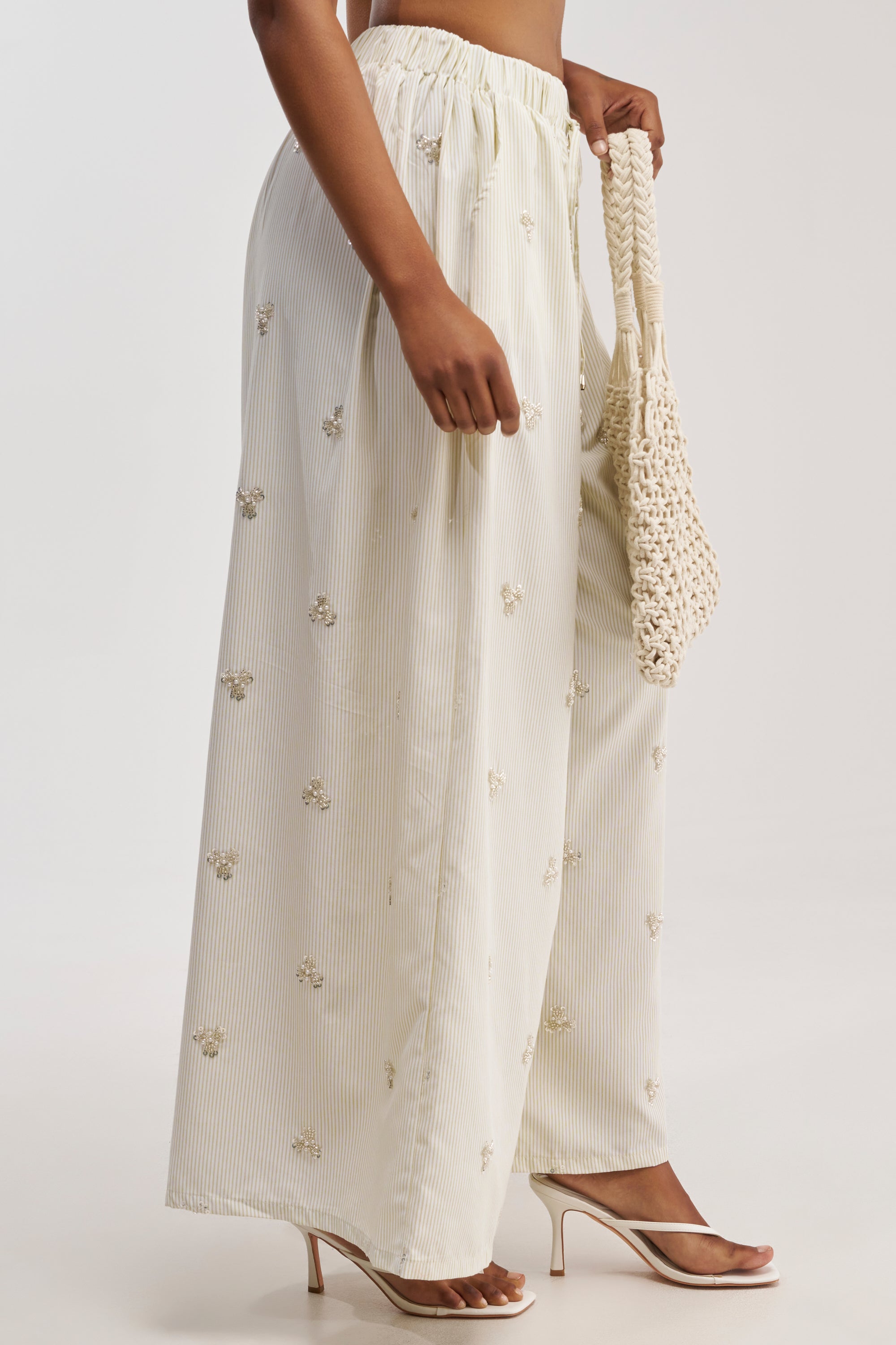 A woman wears the LIGHT AND AIRY POPLIN PANT in cream, featuring a wide-leg silhouette with delicate small embroidered flowers. She carries a woven bag and completes the look with open-toe high heels, set against a plain white background.